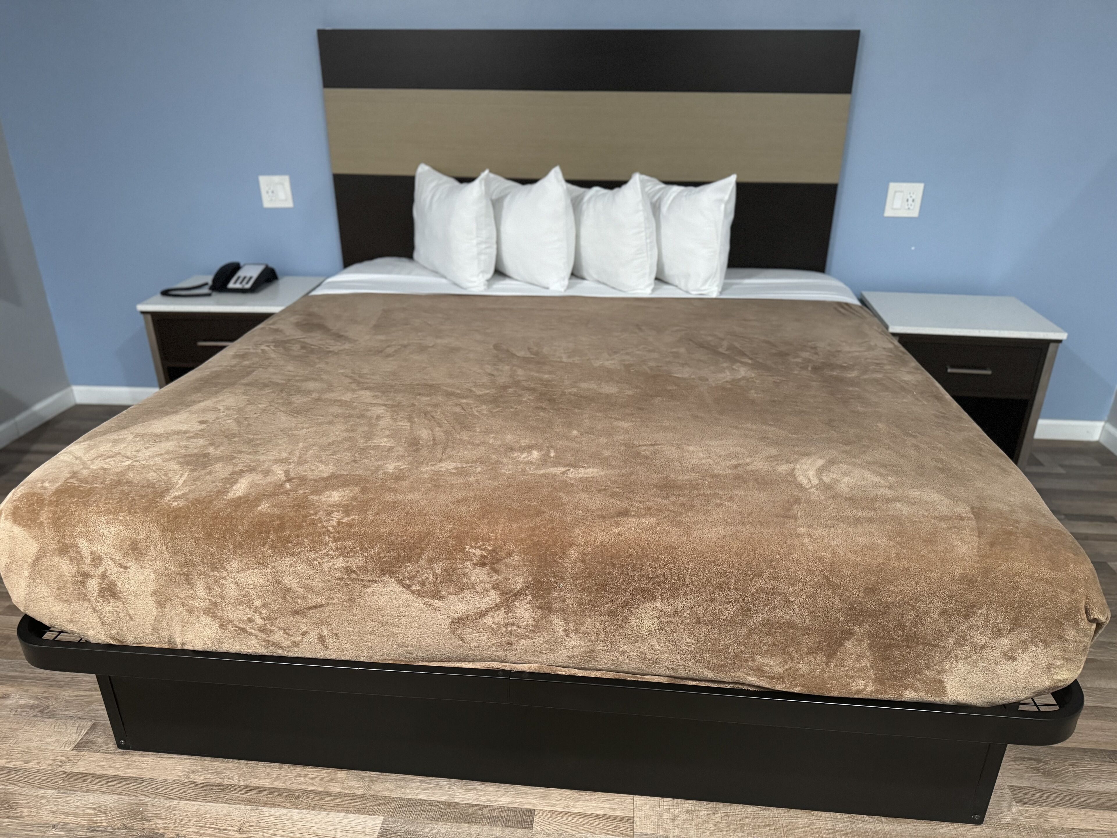 Una cama con cubierta marrón, cabecera negra y almohadas blancas. Dos mesitas de noche, una con un teléfono y una lámpara, y otra con un cajón. Las paredes son de color azul claro y el suelo de madera.