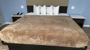 A bed with a brown cover, a black headboard, and white pillows. There are two bedside tables with a phone and a lamp on one, and a drawer on the other. The room has light blue walls and wooden flooring.