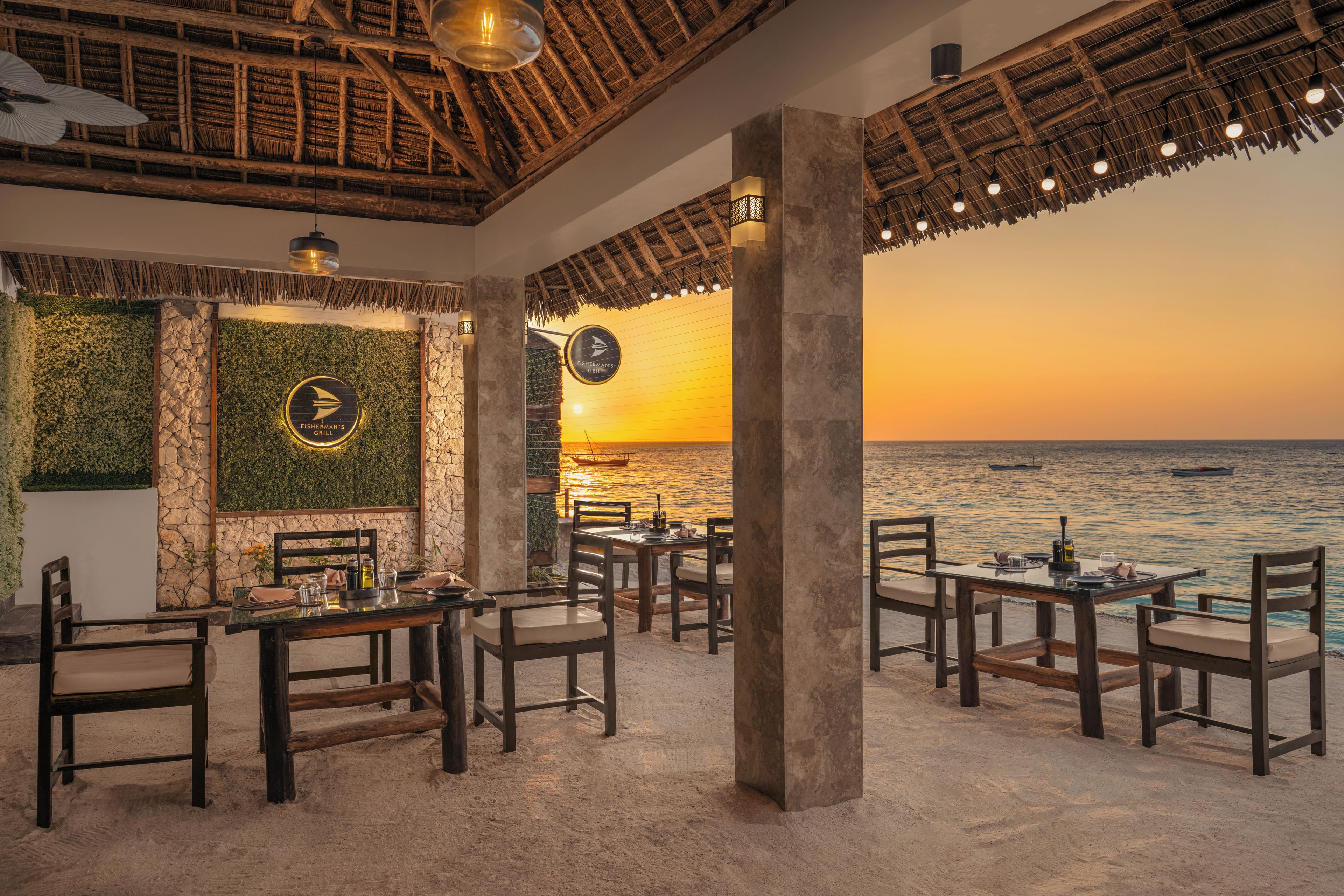 A restaurant with a view of the sunset over the water, featuring tables and chairs, a thatched roof, and a stone wall with greenery.