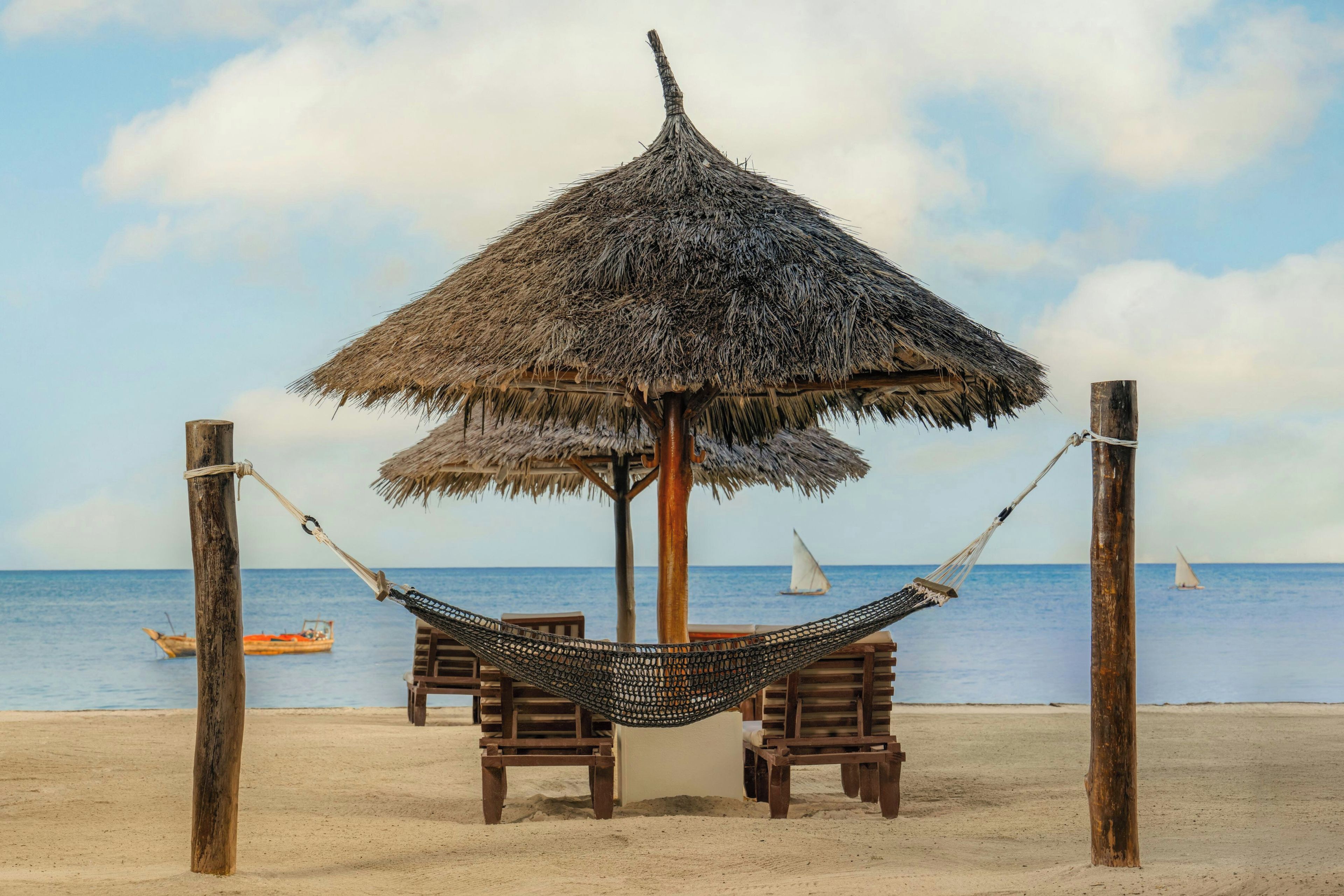 A beach scene with a thatched-roof structure, wooden benches, and a hammock.
