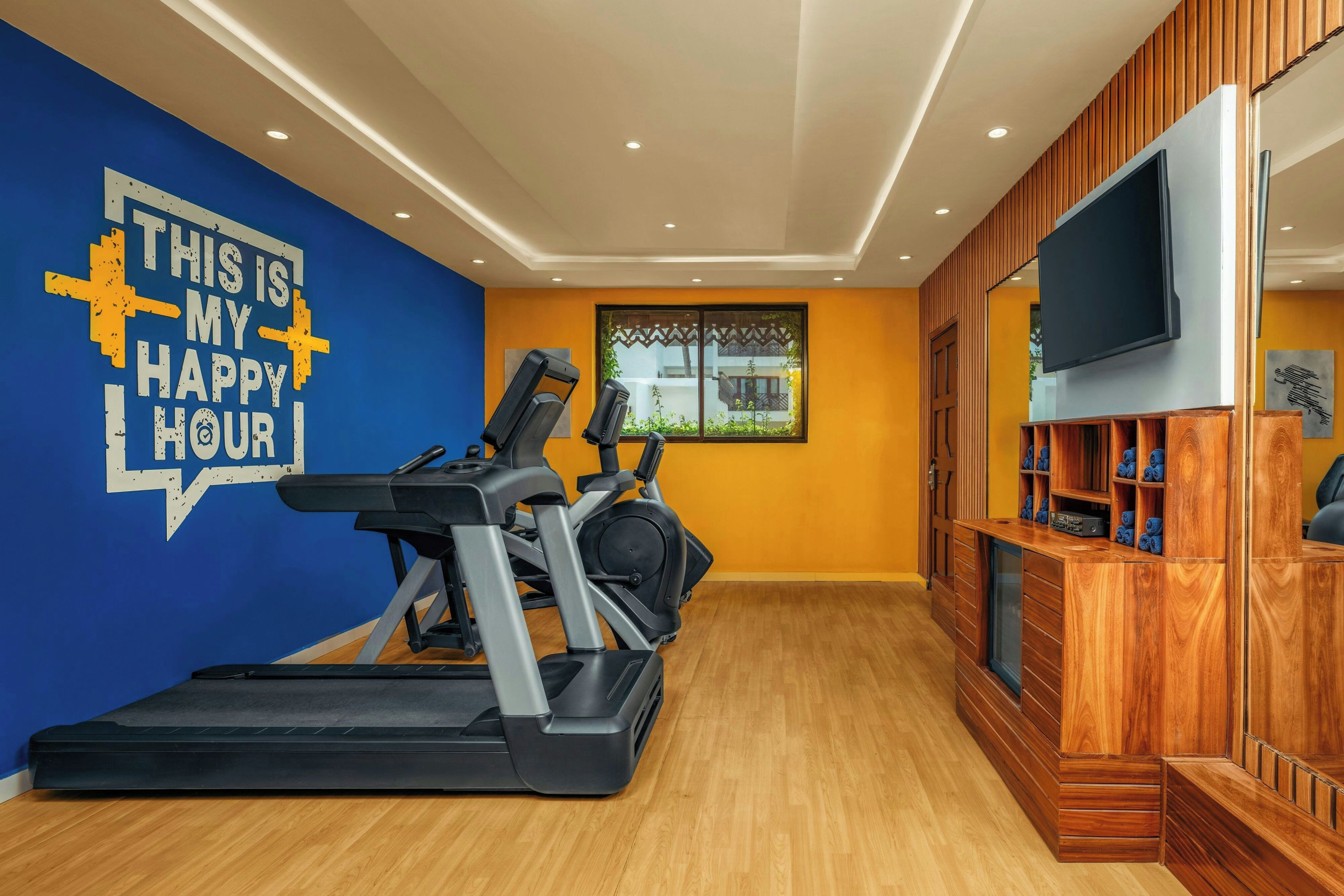 Un gimnasio en casa con cintas de correr, una pared con cita motivacional, una ventana con vistas y un televisor sobre un mueble de madera.