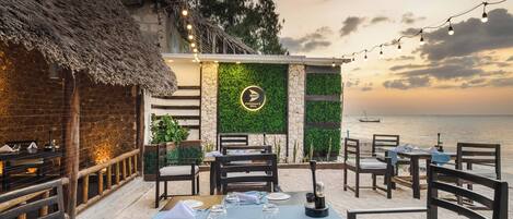 A restaurant with outdoor seating overlooking the sea, featuring a thatched-roof building and a green wall with a logo.