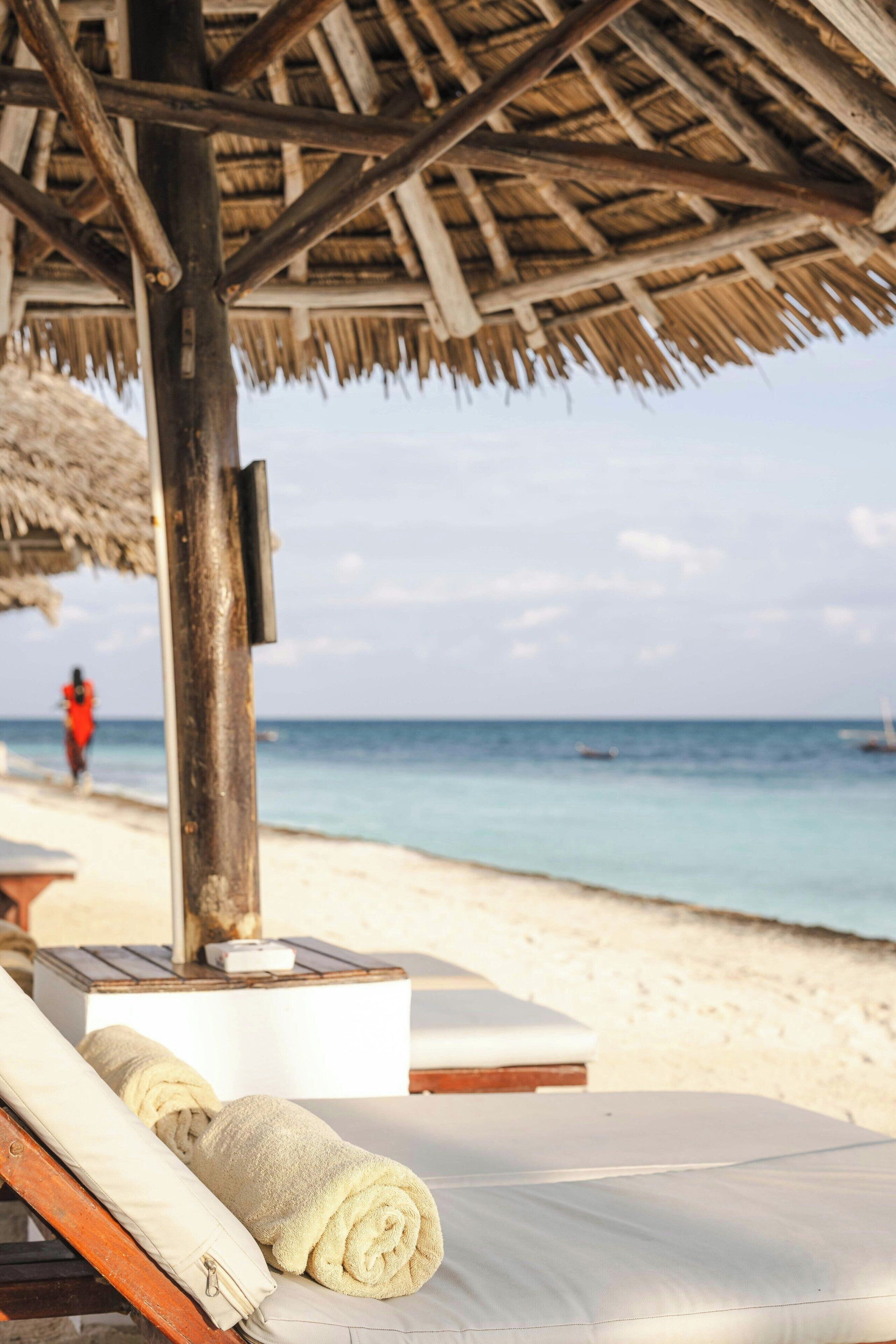 A beachside lounge with a thatched roof, a wooden table, and a towel on a lounge chair.