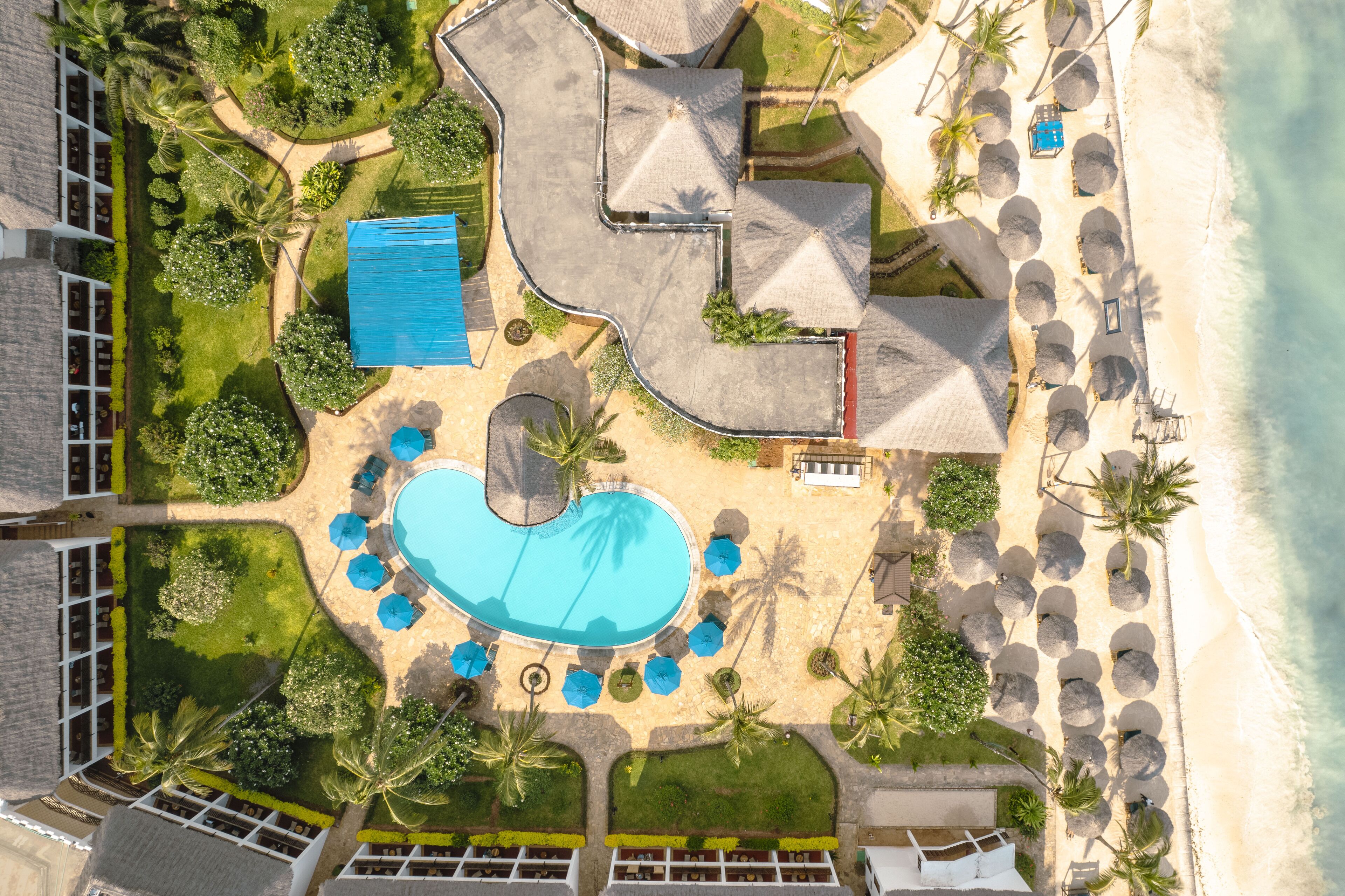An aerial view of a resort with a swimming pool, beachside seating, and palm trees.