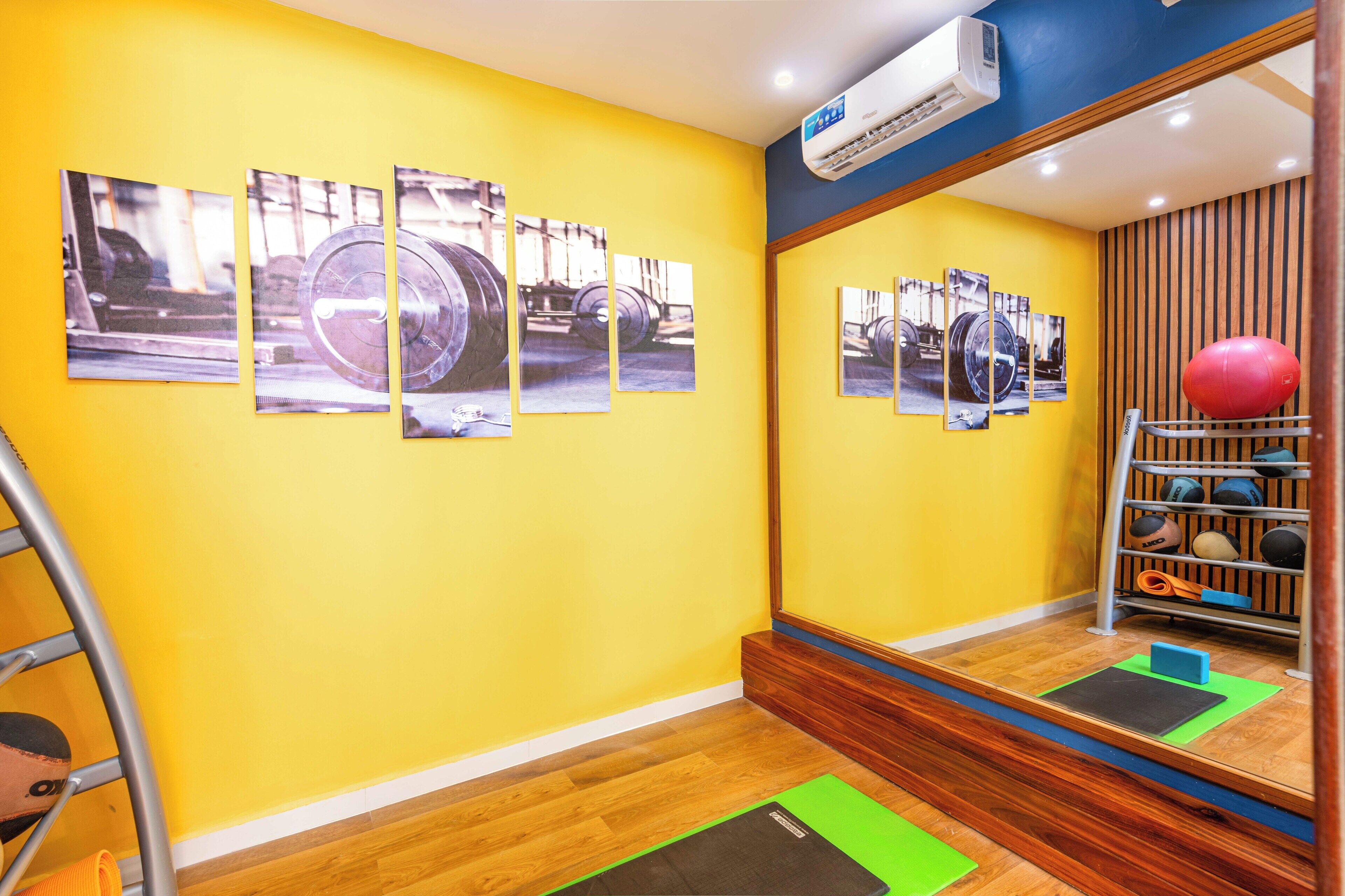 A room with a yellow wall, wooden flooring, a large mirror reflecting exercise equipment, and a series of photographs on the wall.