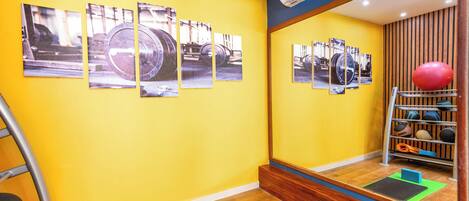 A room with a yellow wall, wooden flooring, a large mirror reflecting exercise equipment, and a series of photographs on the wall.