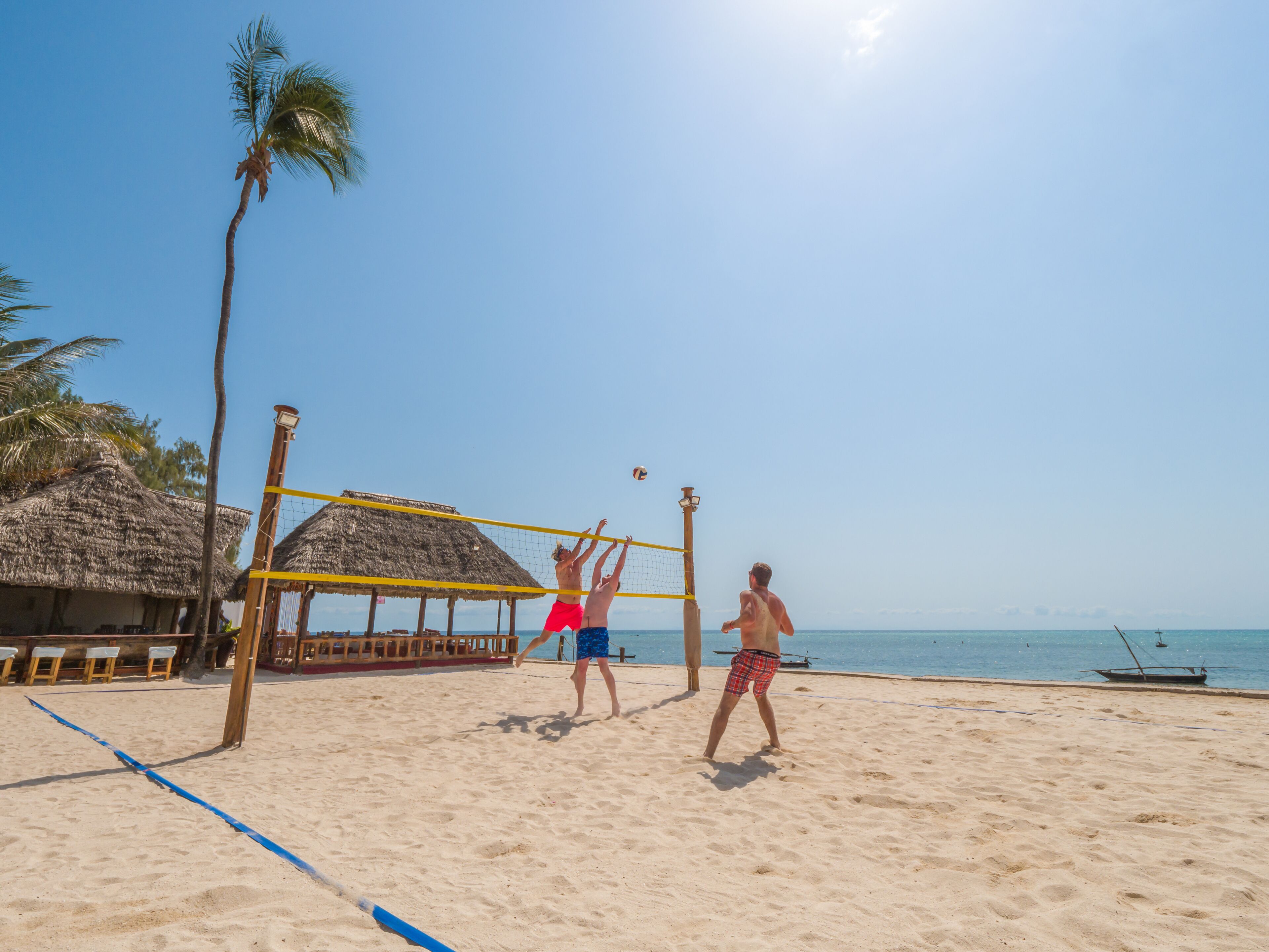 A beach volleyball game on a sandy beach with a thatched-roof hut in the background.