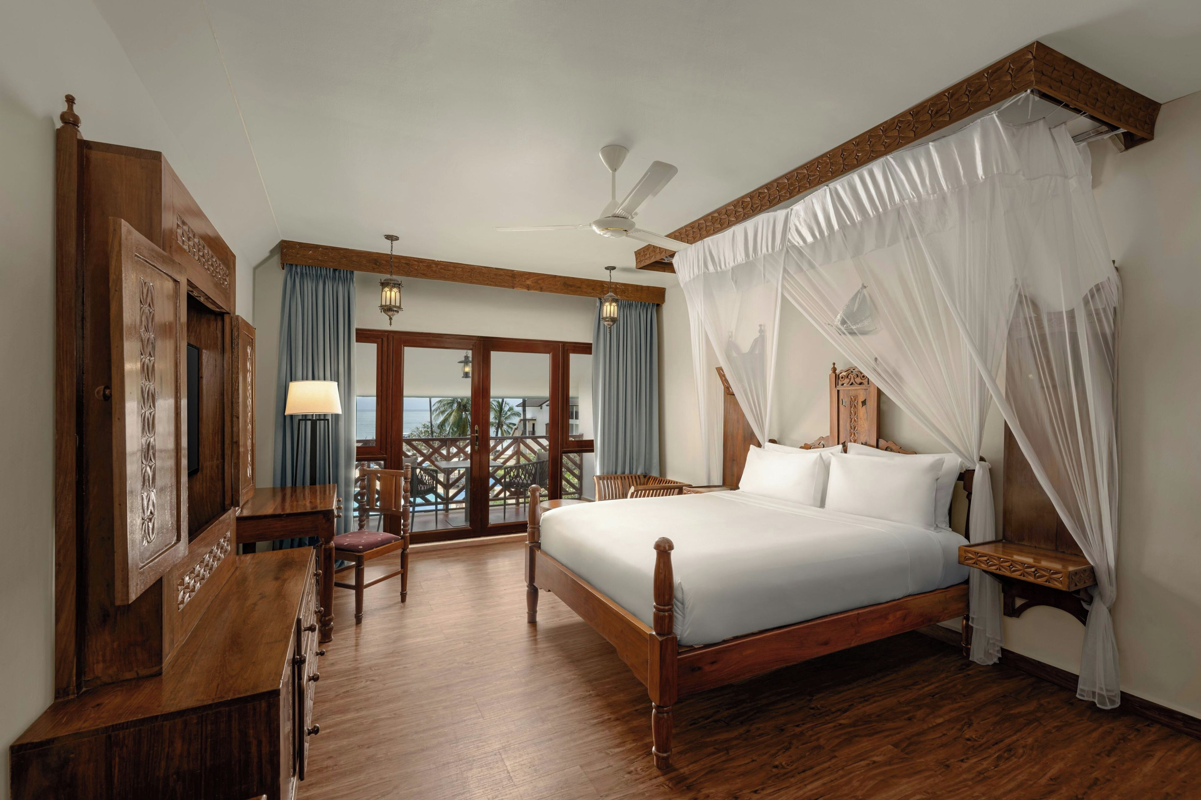 A hotel room with a wooden bed, a desk, a chair, a ceiling fan, and a view of the ocean through the windows.