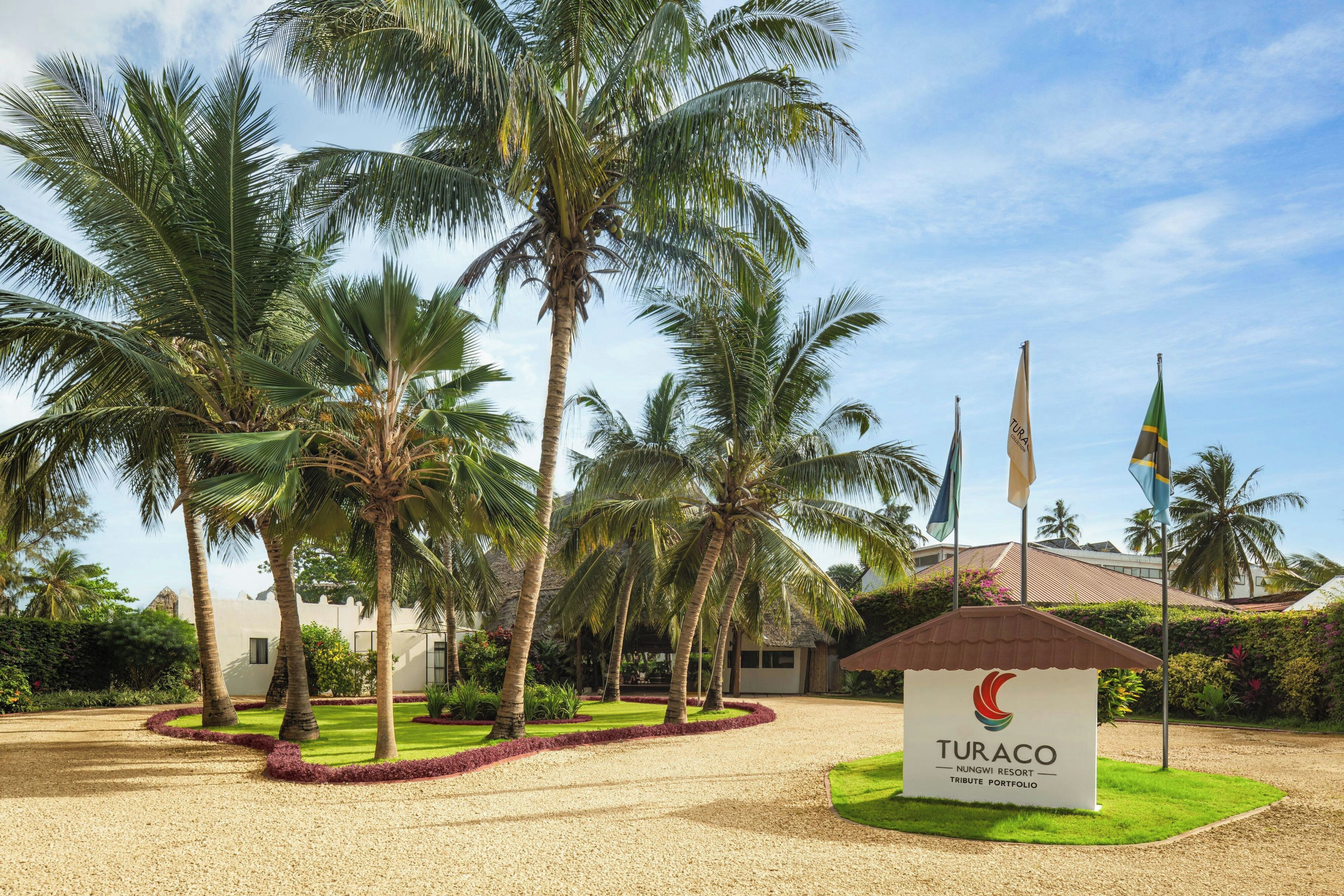Entrada de un resort con palmeras, un cartel y banderas.