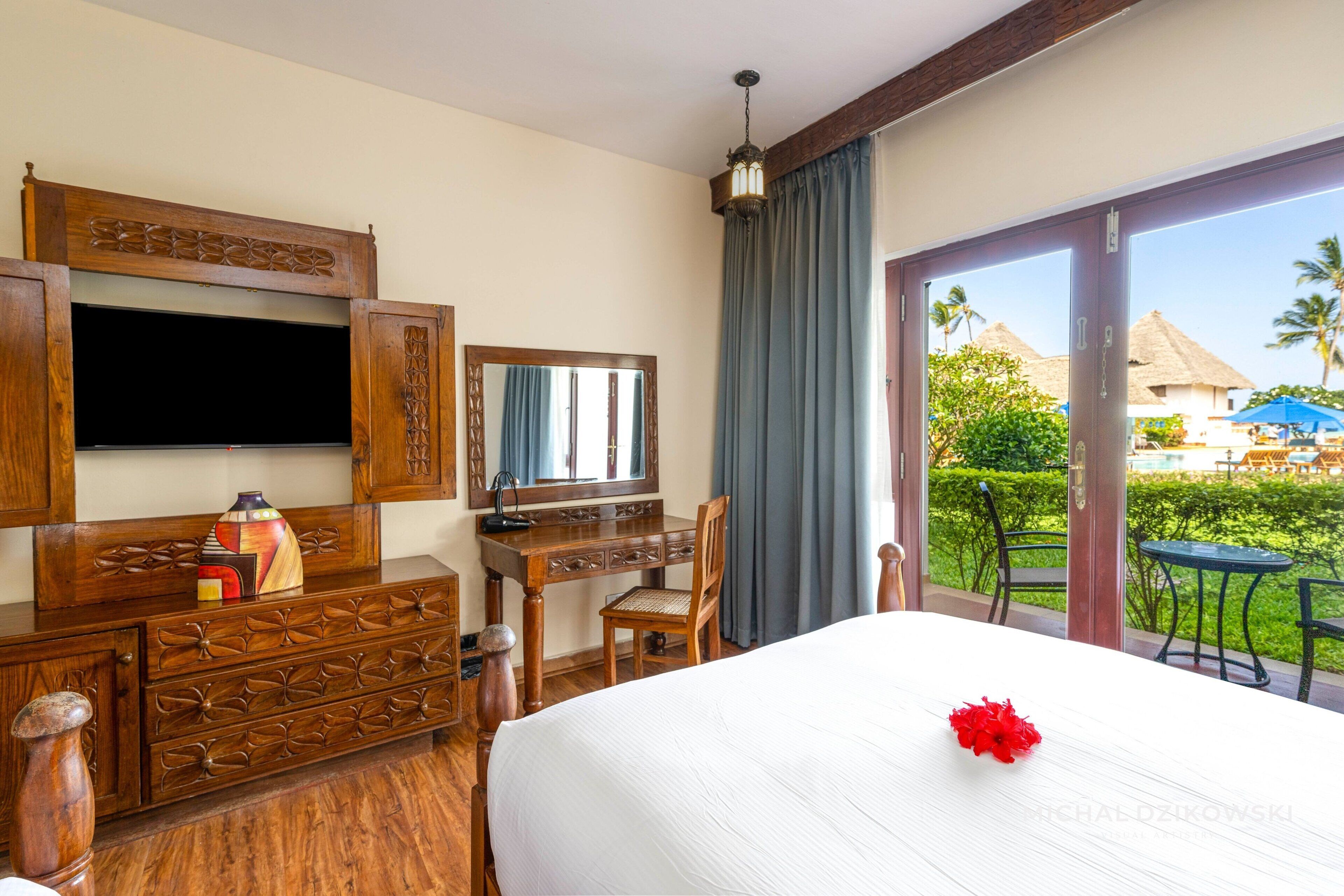 A hotel room with a bed, a desk, a chair, a TV, and a balcony with a view of a resort.