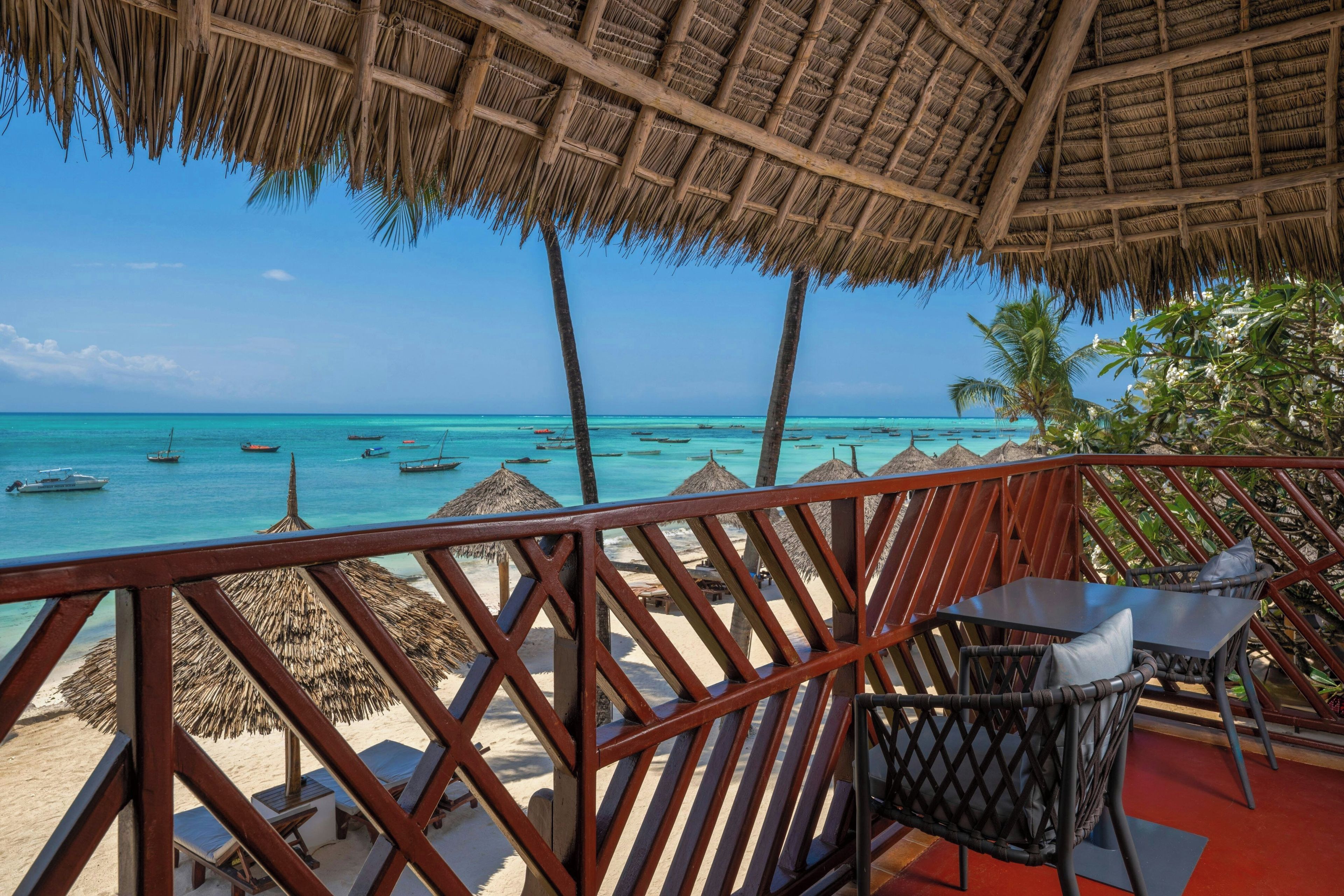 A beachside view with a thatched roof structure, a wooden railing, and a clear view of the ocean with boats.