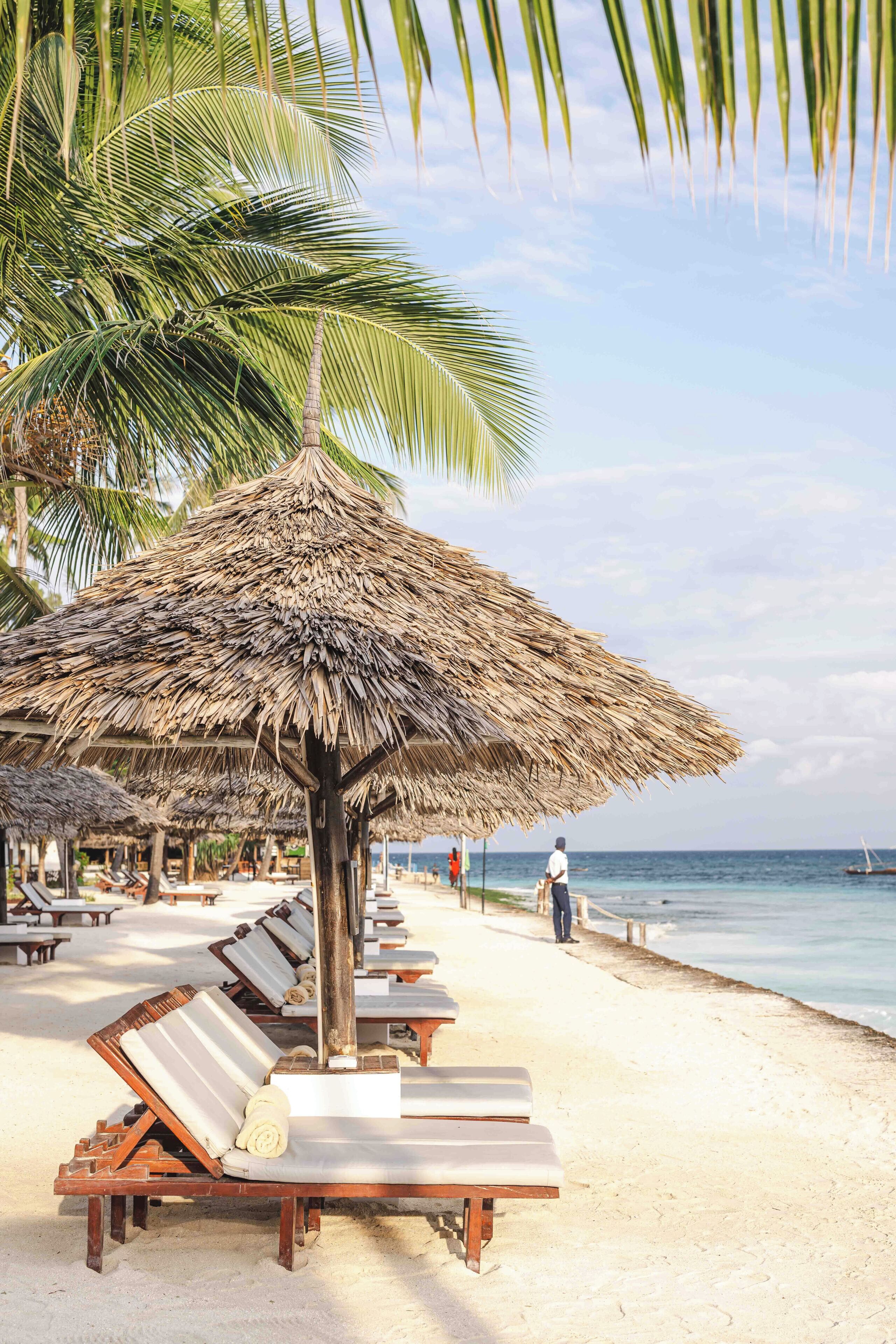 A beach with thatched umbrellas, lounge chairs, and palm trees.