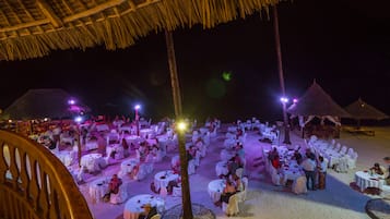 A beachside dining area with round tables and chairs, surrounded by thatched umbrellas, under a wooden roof structure.