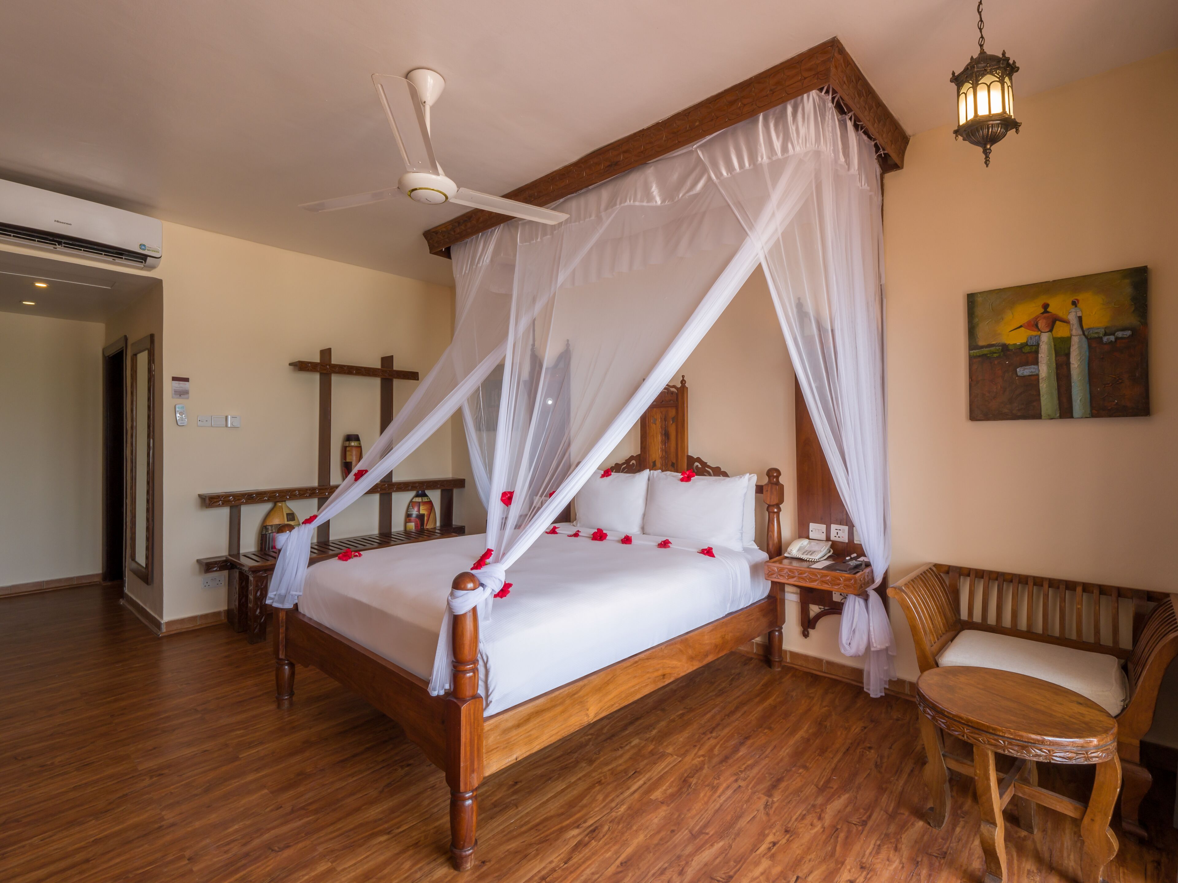 A four-poster bed with a canopy, a wooden floor, a ceiling fan, a bedside table with a lamp, a chair, and a painting on the wall.