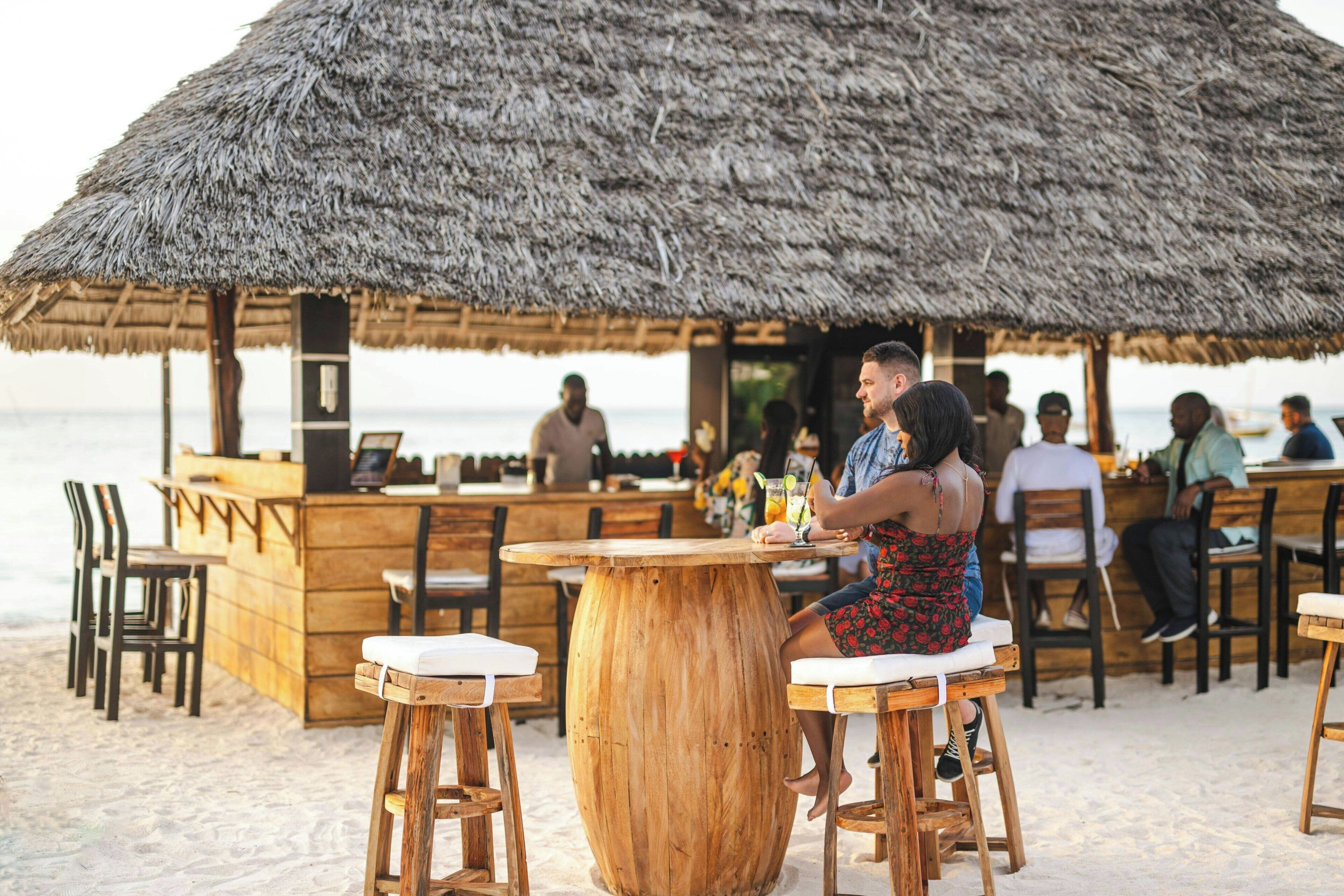 A beachside bar with wooden tables and chairs, a thatched roof, and a view of the ocean.