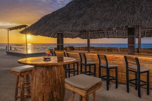 Un bar frente a la playa con mobiliario de madera y techo de paja.