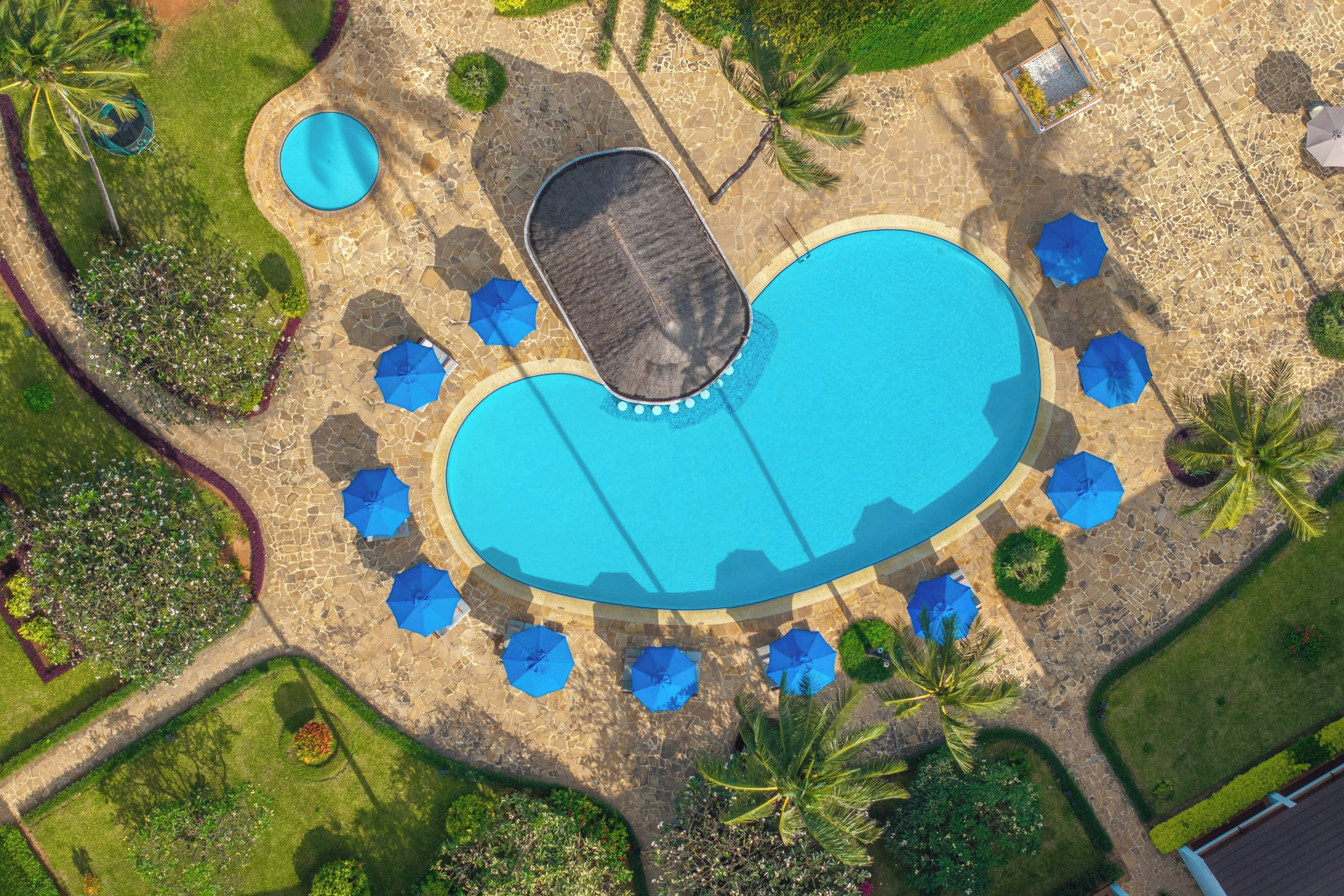 An aerial view of a resort with a large central swimming pool, surrounded by palm trees and blue umbrellas.