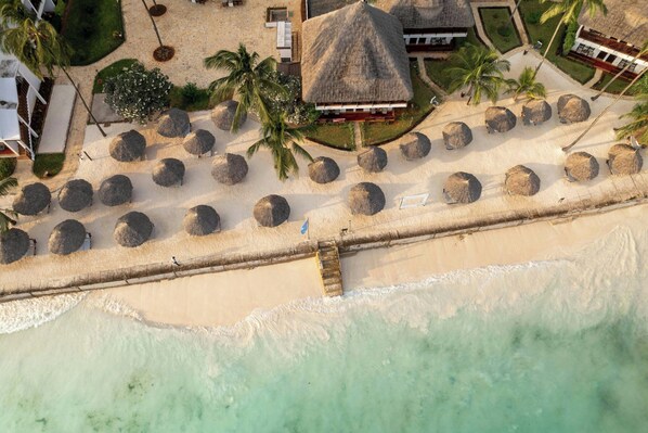 An aerial view of a beach resort with thatched-roof structures, palm trees, and a sandy beach.