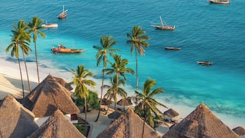 A beach with thatched-roof huts, palm trees, and boats on the water.