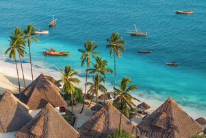 Una playa con chozas de techo de paja, palmeras y barcos en el agua.