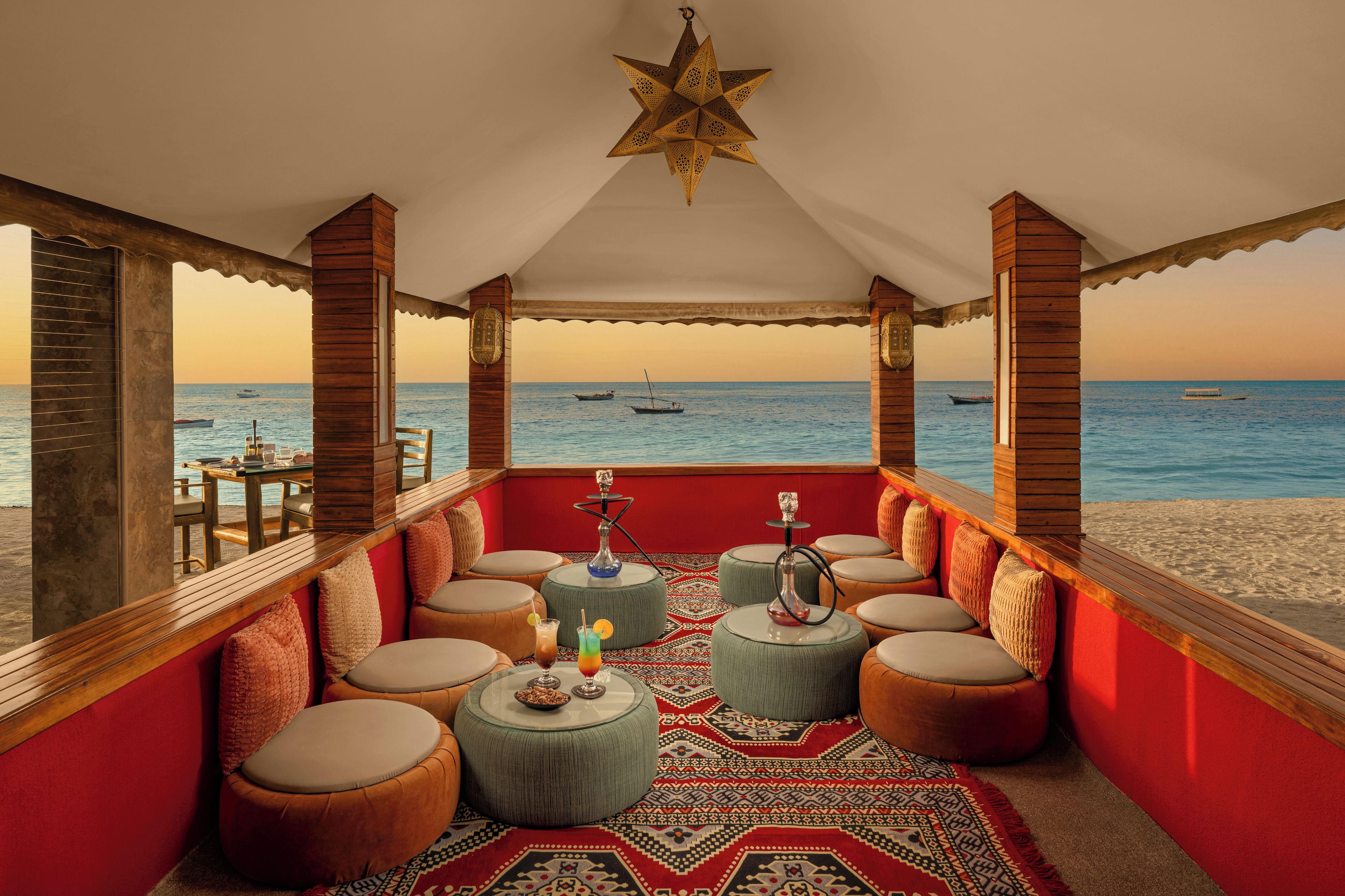 A covered outdoor seating area with a view of the sea, featuring round tables and ottomans, a star-shaped ceiling light, and a patterned rug.