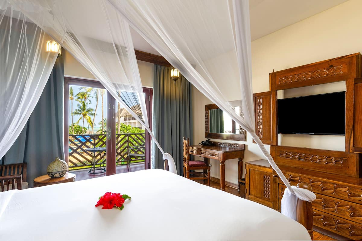A hotel room with a bed, a desk, a television, and a balcony with a view of palm trees.