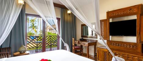 A hotel room with a bed, a desk, a television, and a balcony with a view of palm trees.
