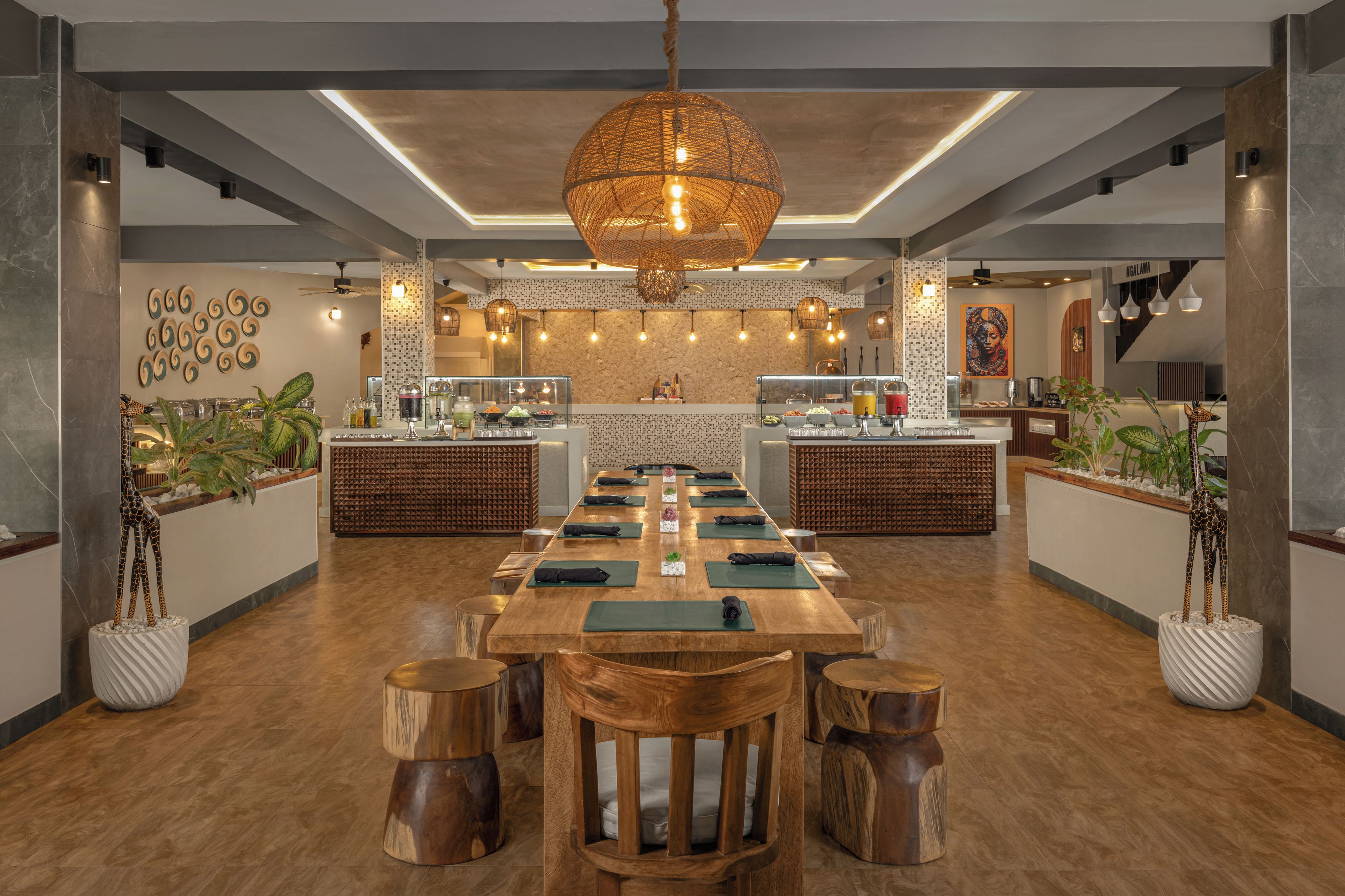 A modern restaurant with a long wooden table, cushioned chairs, and a central hanging light fixture.