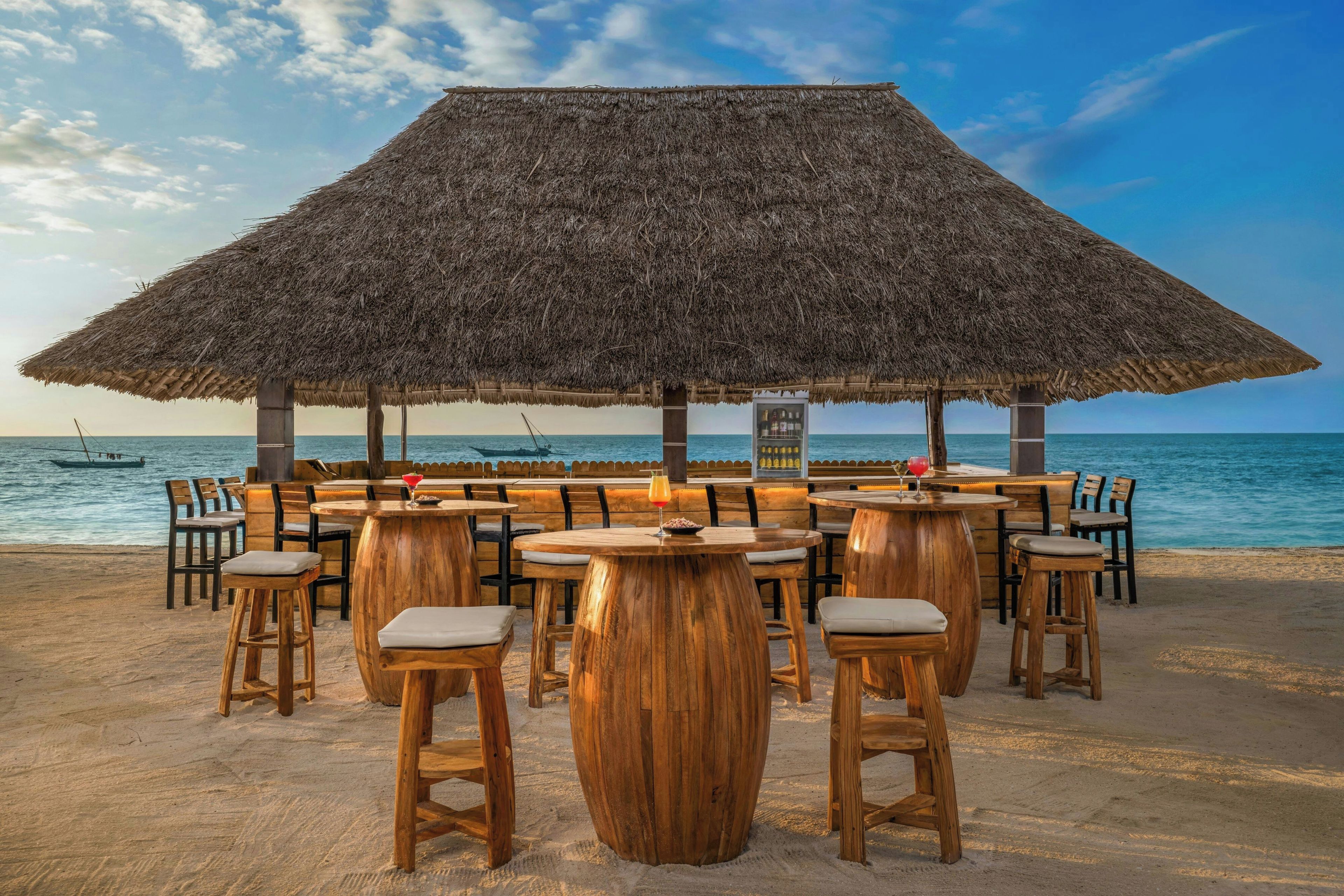 Eine Strandbar mit einem Reetdach, Holztischen und -stühlen sowie Blick auf den Ozean.