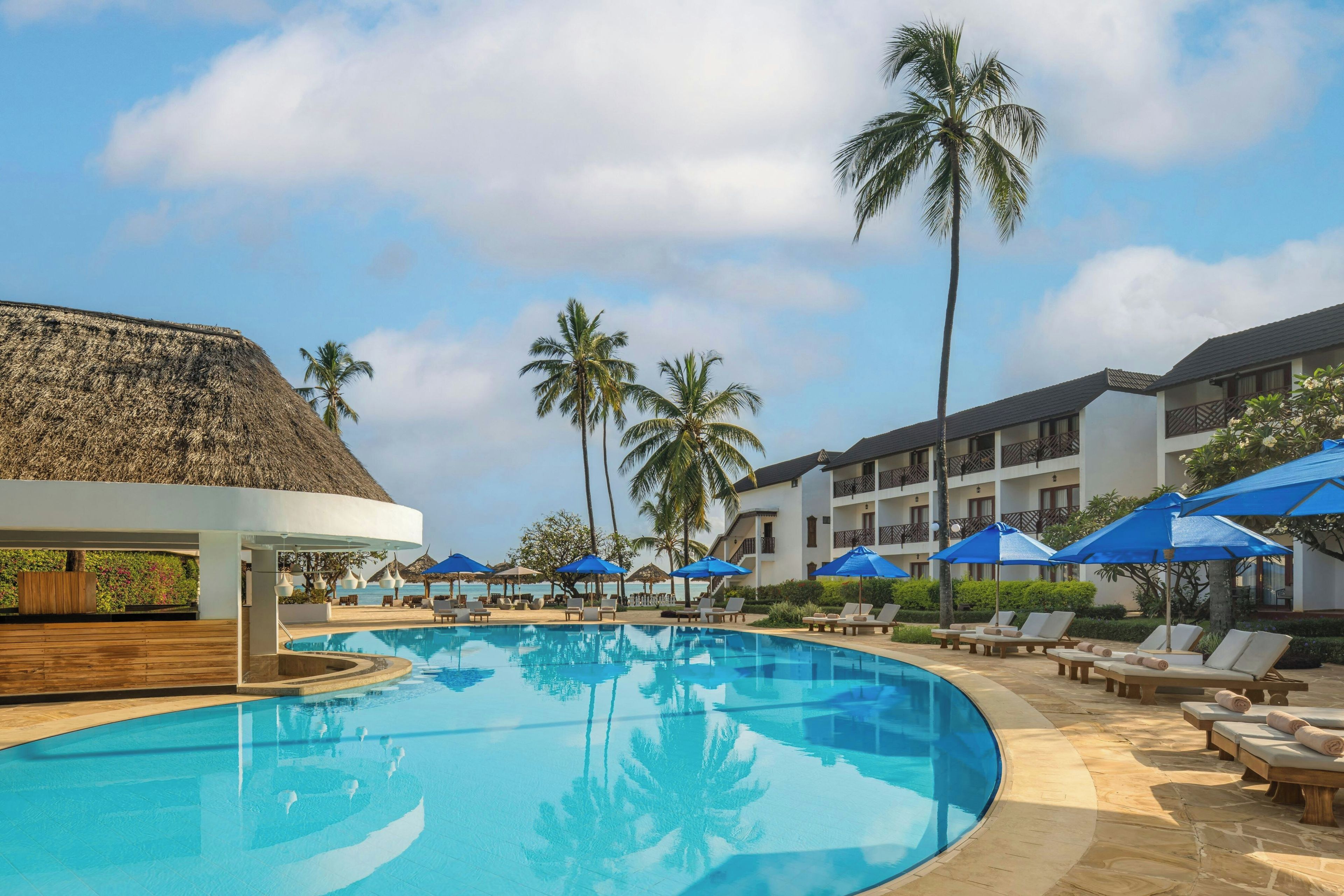 A resort pool area with lounge chairs and umbrellas, surrounded by palm trees and a building with balconies.