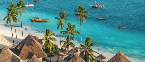 A beach with thatched-roof huts, palm trees, and boats on the water.