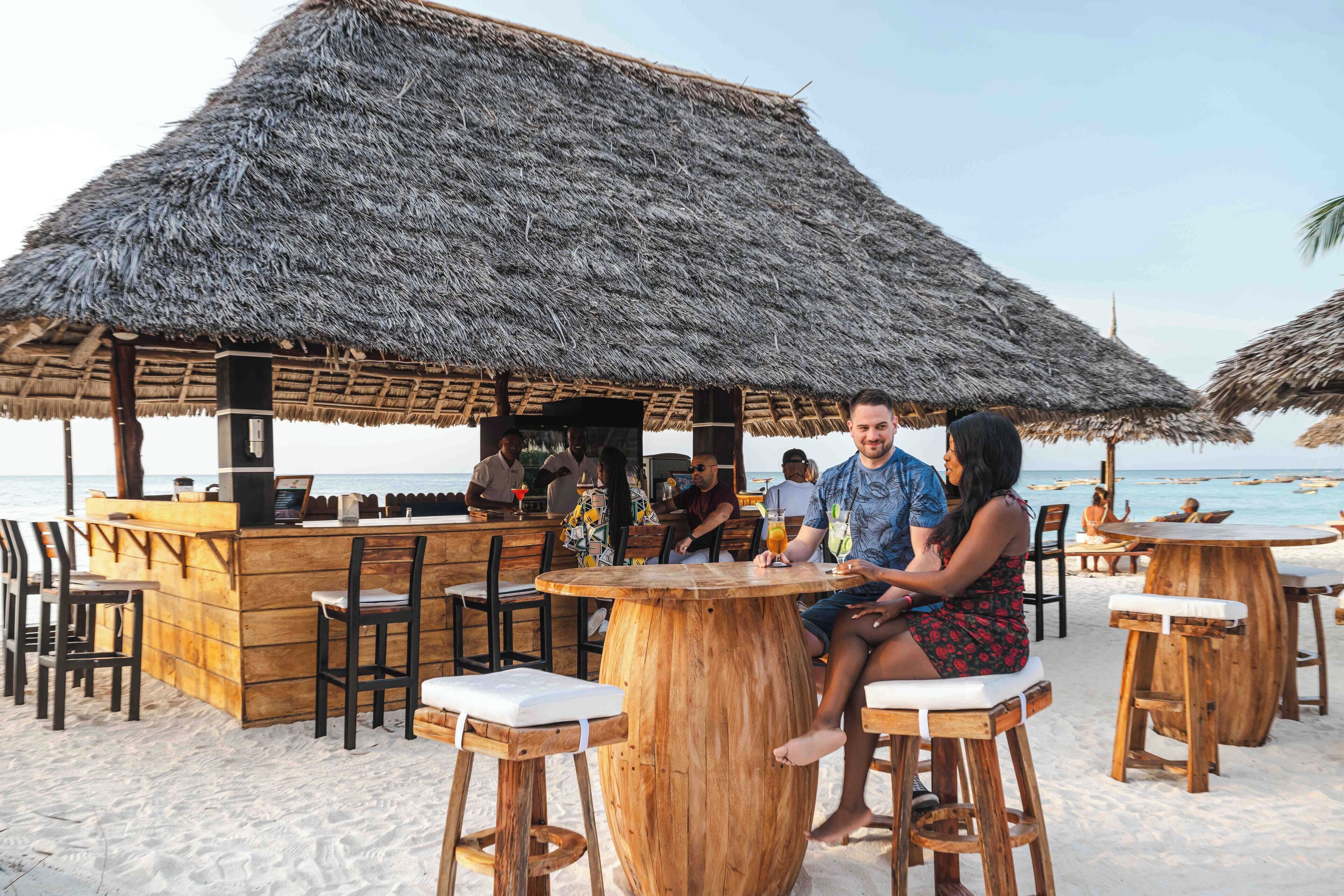 A beachside bar with wooden stools and thatched umbrellas.