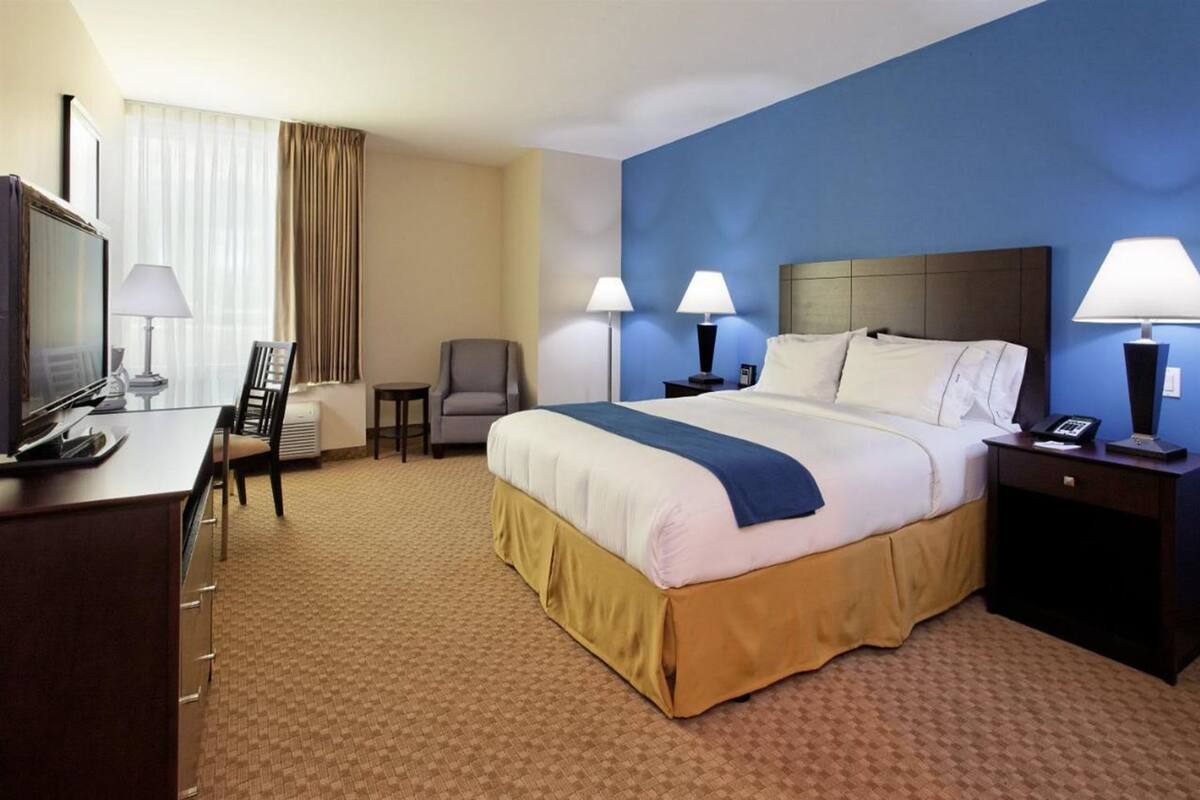 Une chambre d’hôtel avec un grand lit, deux tables de chevet avec des lampes, un bureau avec une chaise, une télévision et une fenêtre avec des rideaux.