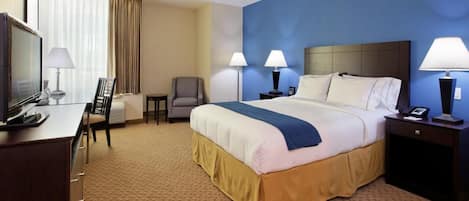 A hotel room with a large bed, two bedside tables with lamps, a desk with a chair, a TV, and a window with curtains.