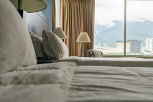 Quarto de hotel com cama, duas luminárias de cabeceira, uma cadeira e vista para montanhas e edifícios através de amplas janelas.