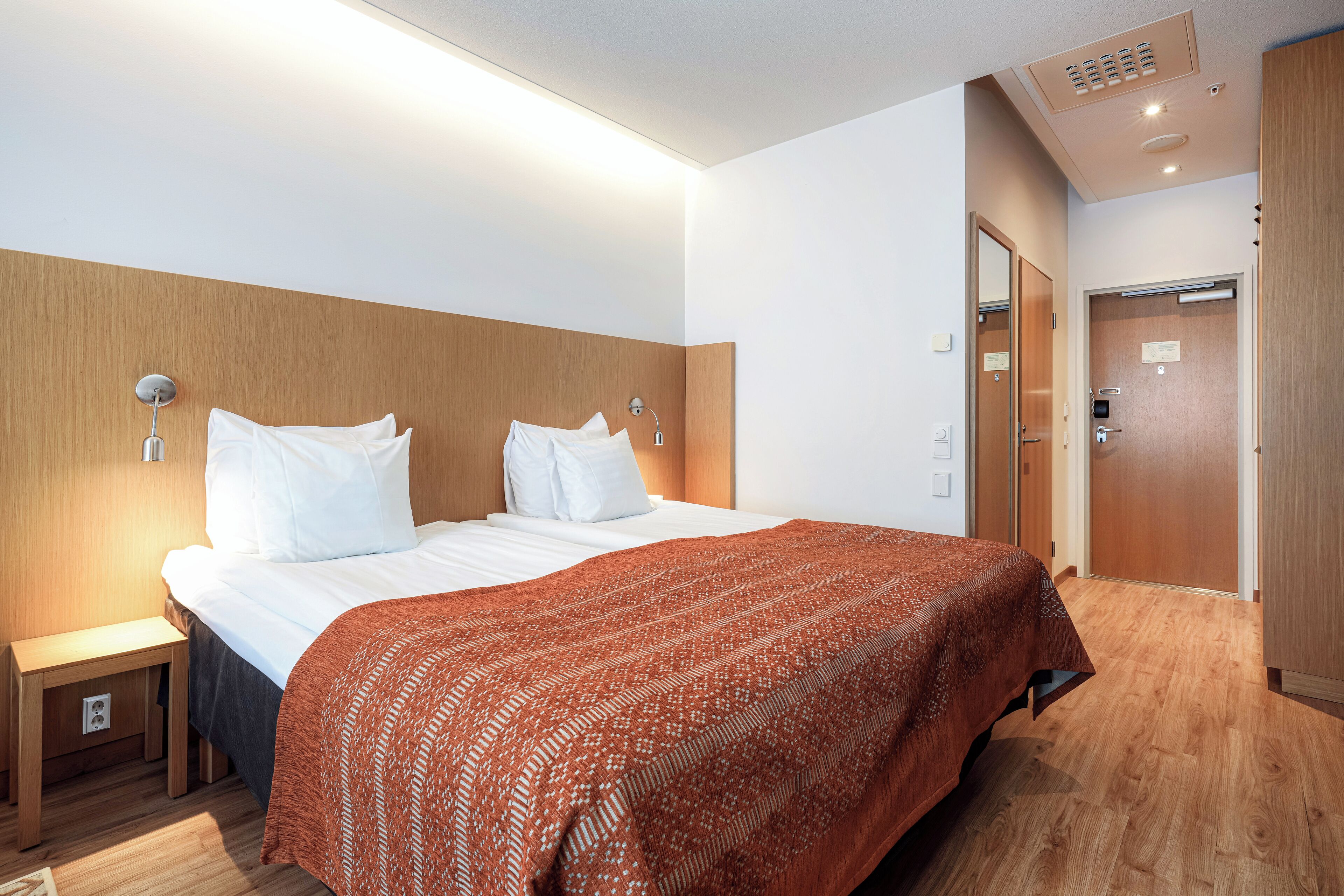 A hotel room with a large bed, wooden headboard, two bedside tables, a nightstand, and a wooden floor.