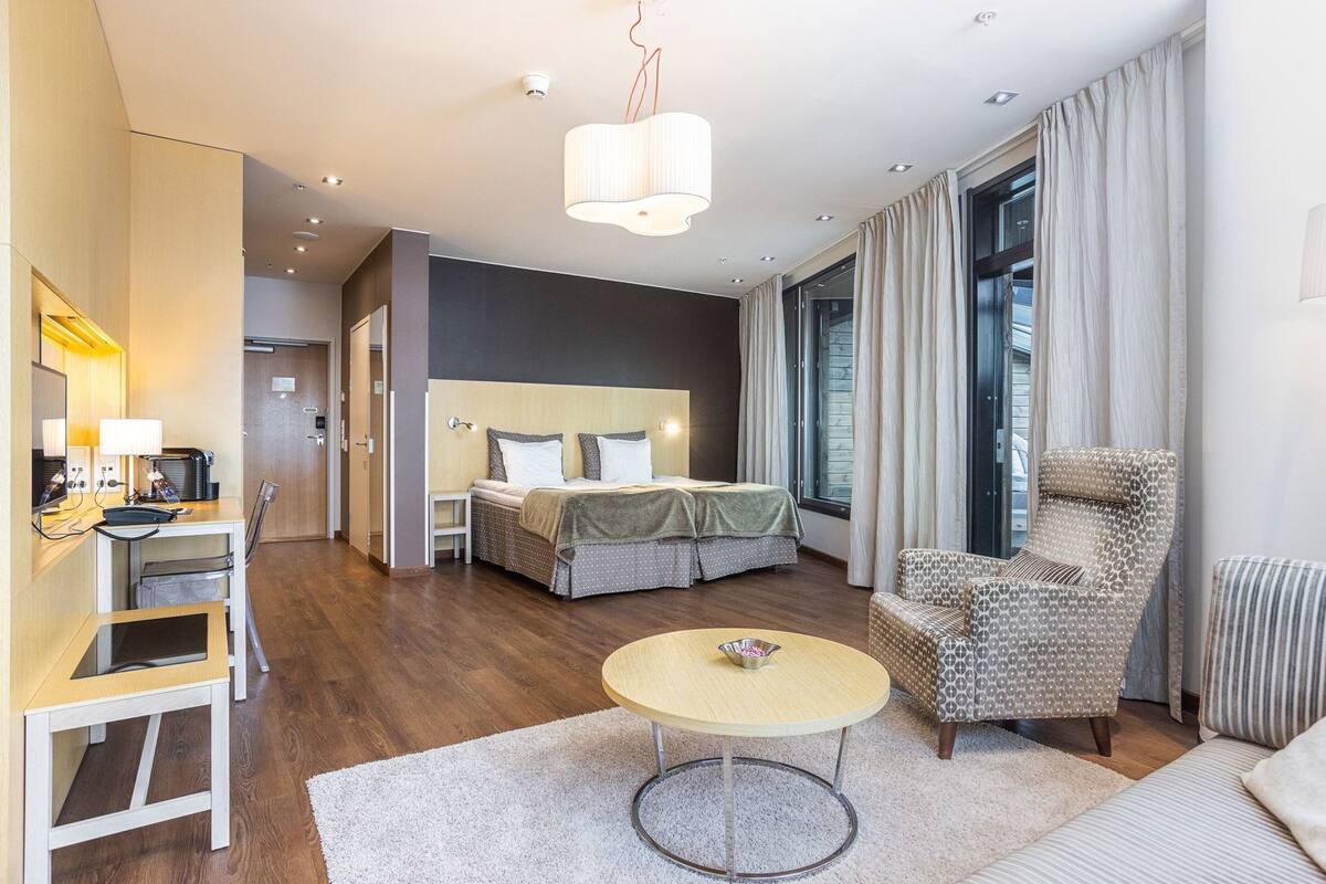 Una habitación de hotel moderna con una cama, un escritorio con silla, una mesa redonda, un sillón estampado y un ventanal con cortinas.
