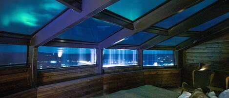 A room with a bed, a window, and a view of the city at night.