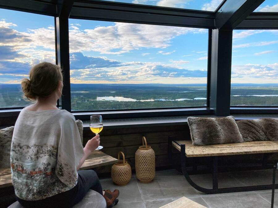 Una persona seduta vicino a una finestra con vista su un lago e una foresta, che tiene in mano un bicchiere di vino.