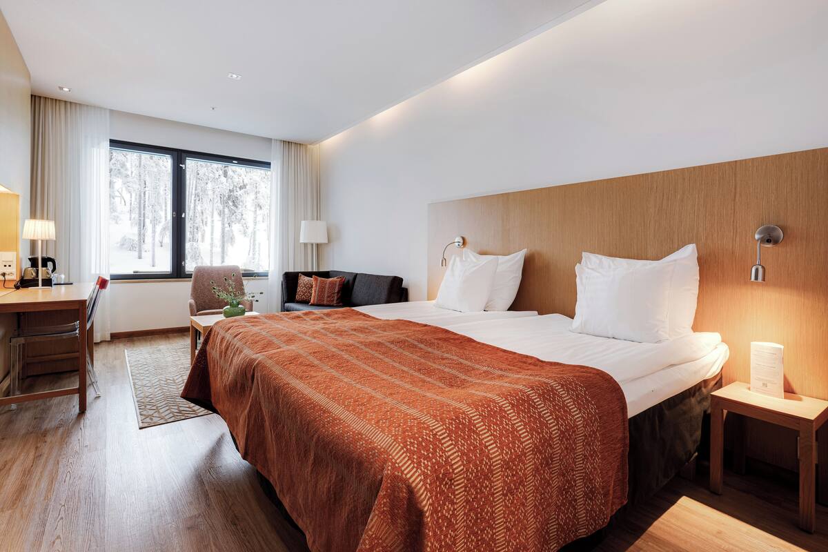 Habitación de hotel con una cama grande, un escritorio con silla, una mesita pequeña y una ventana con cortinas.