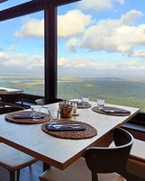 Mesa de comedor dispuesta con individuales, cubiertos y vasos, con vista a un paisaje boscoso.