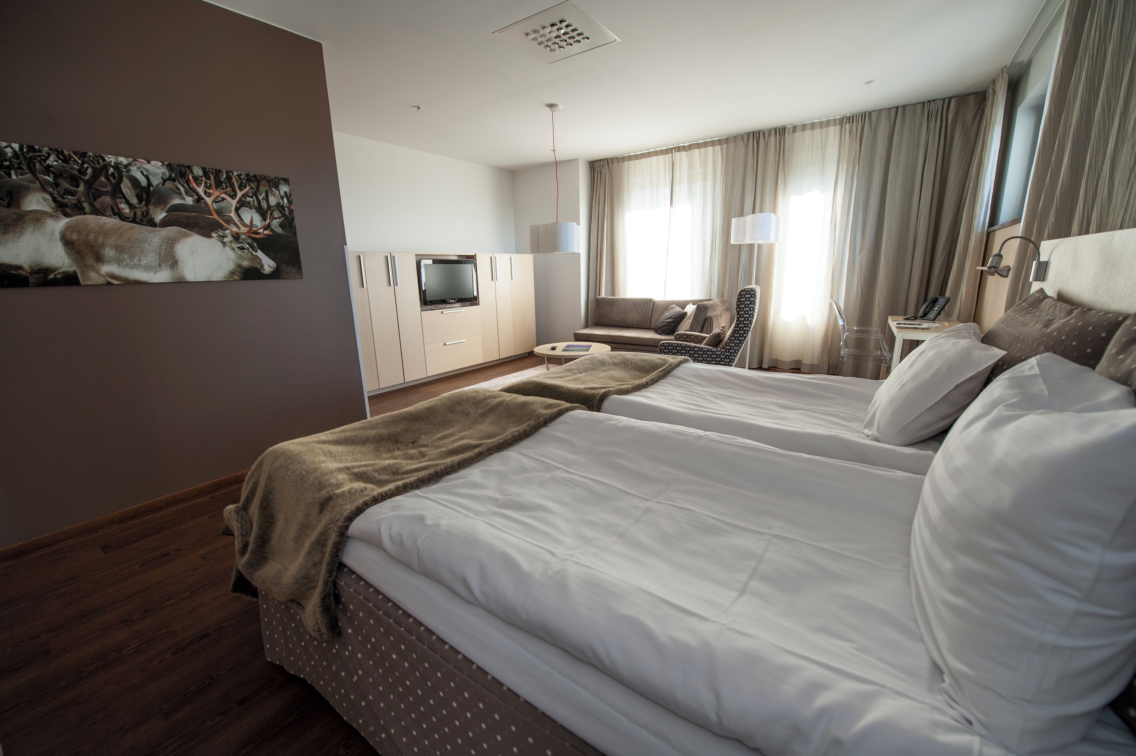 Ett hotellrum med två sängar, en soffa, en tv, ett skrivbord och en tavla med ett motiv av rådjur på väggen.