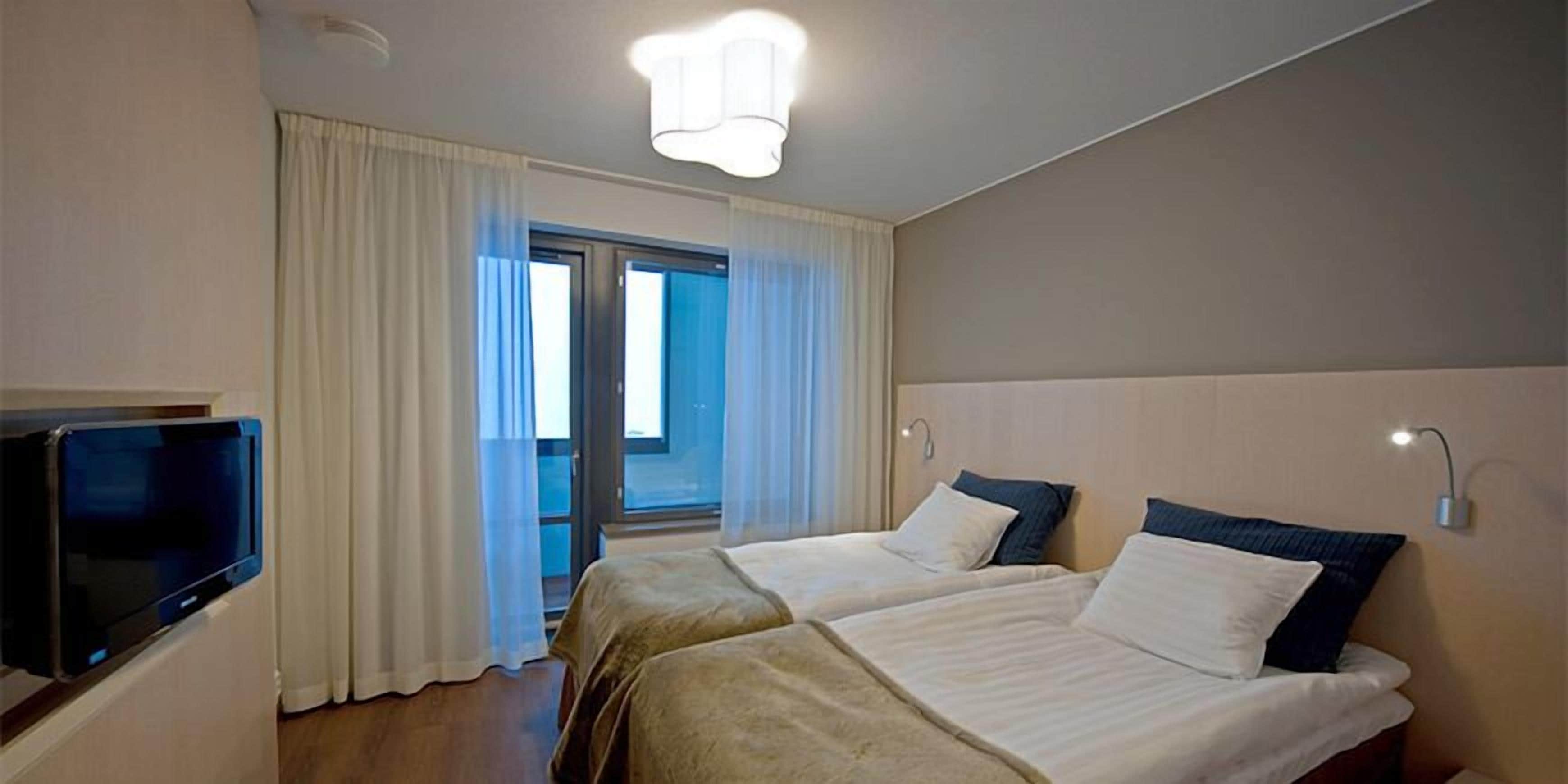 A hotel room with two beds, a television, a ceiling light, and a view of the outdoors through a glass door.