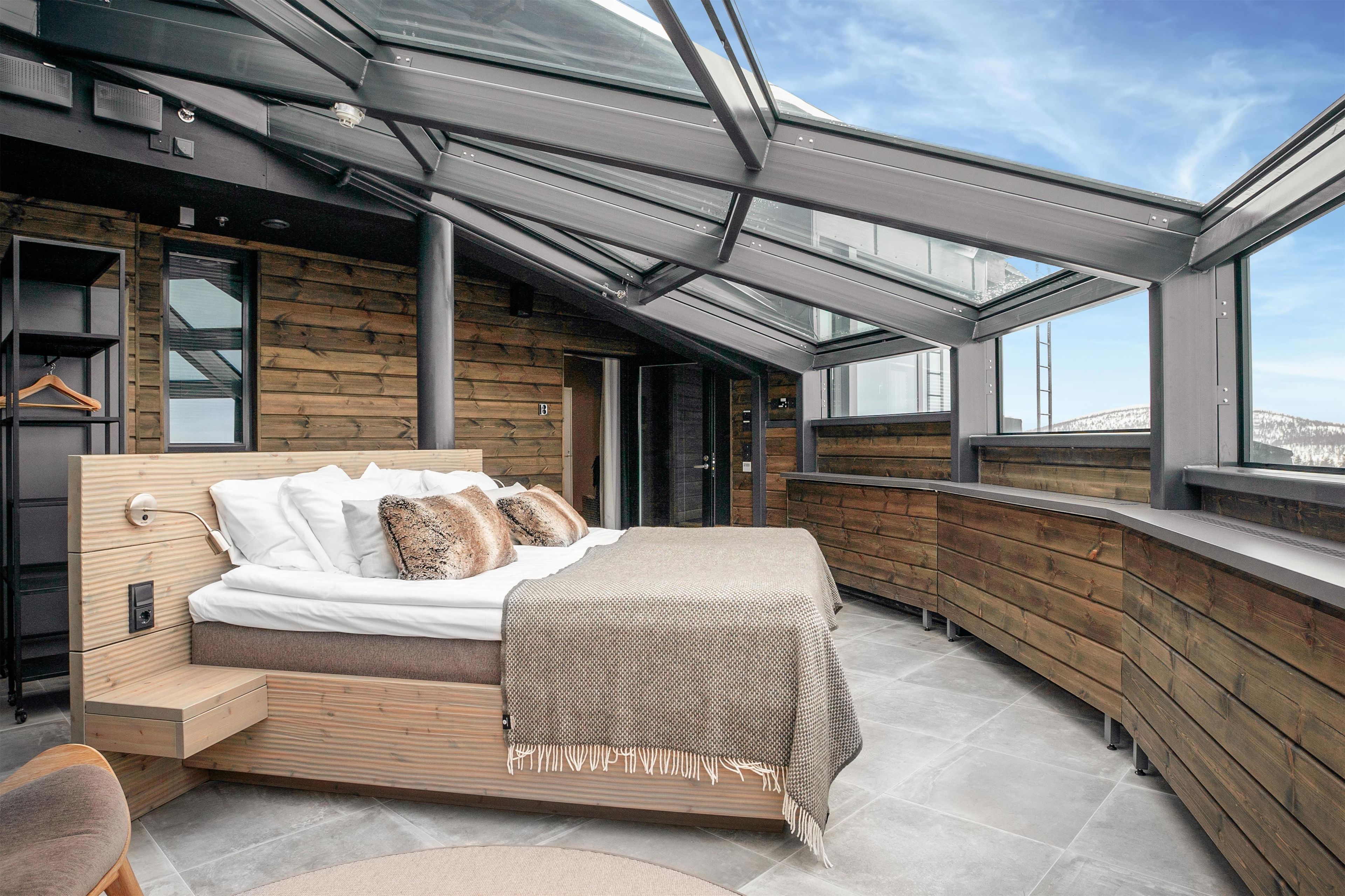 A modern bedroom with a wooden headboard, a bed with white bedding, a bench, and a view of the city through large windows.