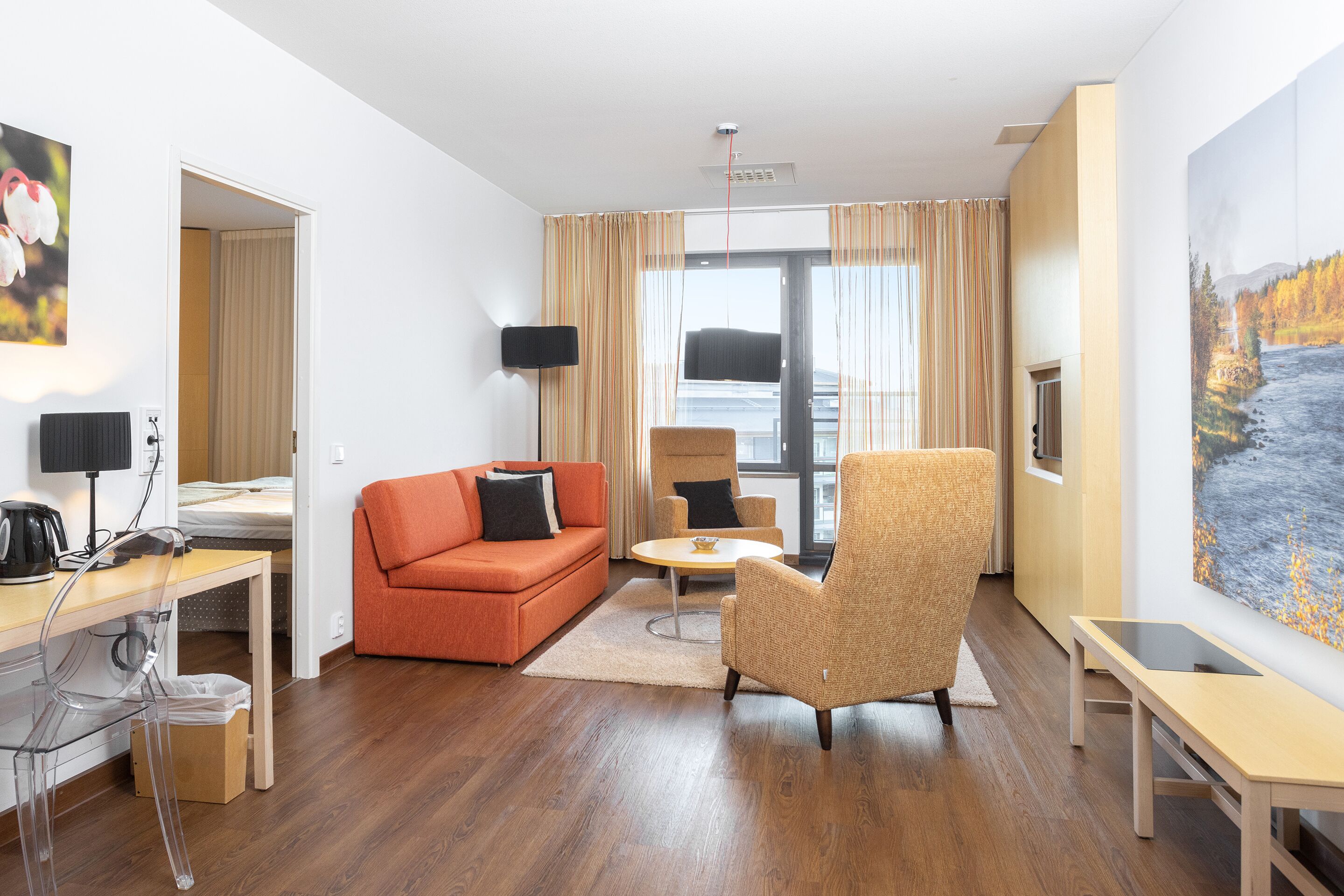 A modern hotel room with a wooden floor, orange sofa, clear plastic chair, and a large painting of a river.