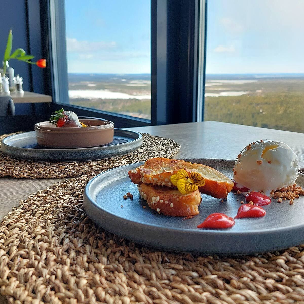 En desserttallerken med pochert æg, en skive brød og en lille skål med sauce, placeret på en vævet underlag med et naturskønt vue i baggrunden.