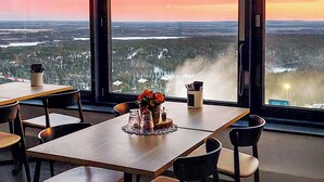 A dining area with a table set for four, large windows, and a view of a cityscape at sunset. - Hotel Levi Panorama (Kittila)
