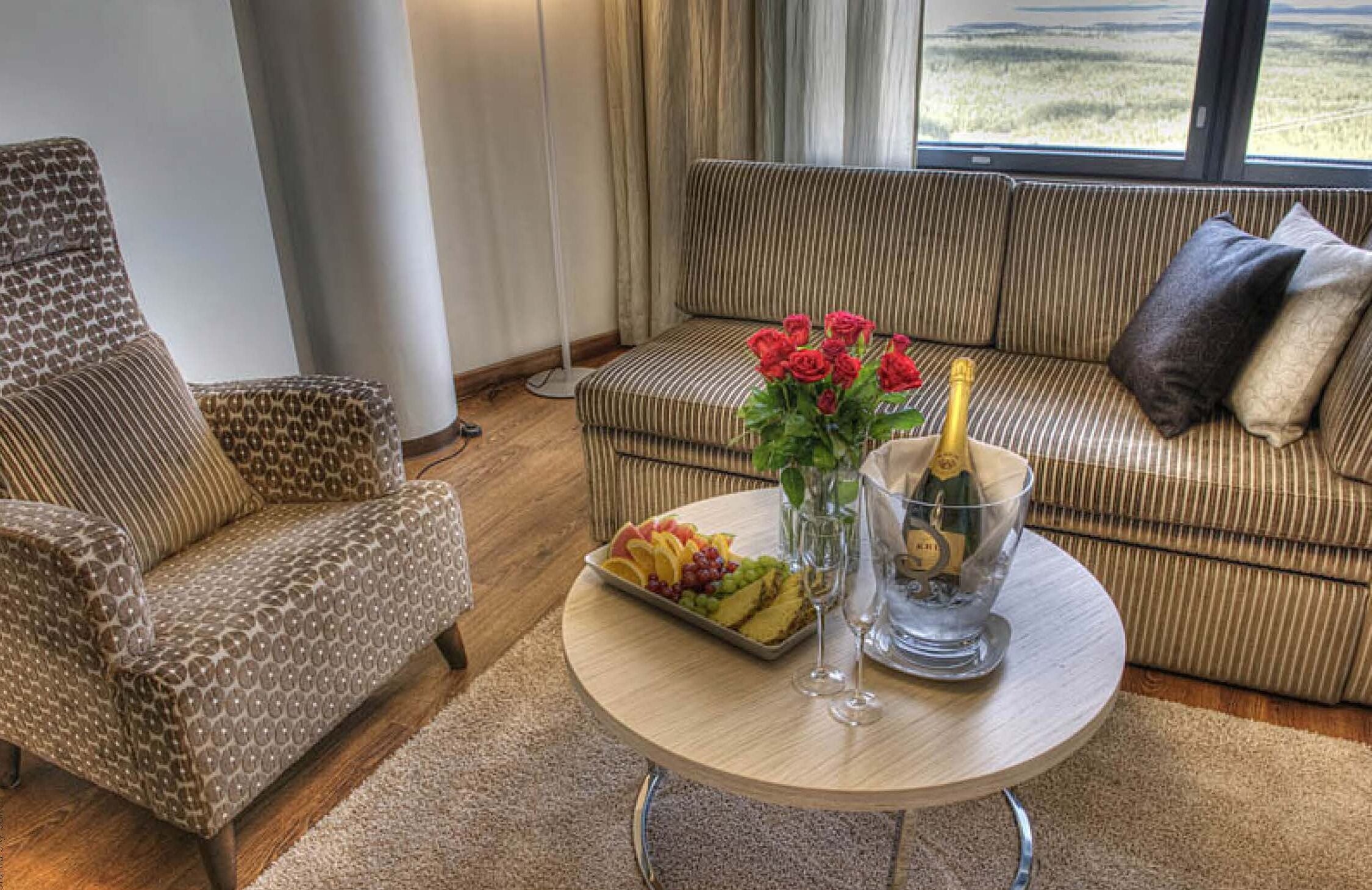 Un salon avec un canapé à rayures, un fauteuil à motifs, une table basse ronde sur laquelle sont disposés des fruits et des boissons, et une vue sur un paysage grâce à de grandes fenêtres.