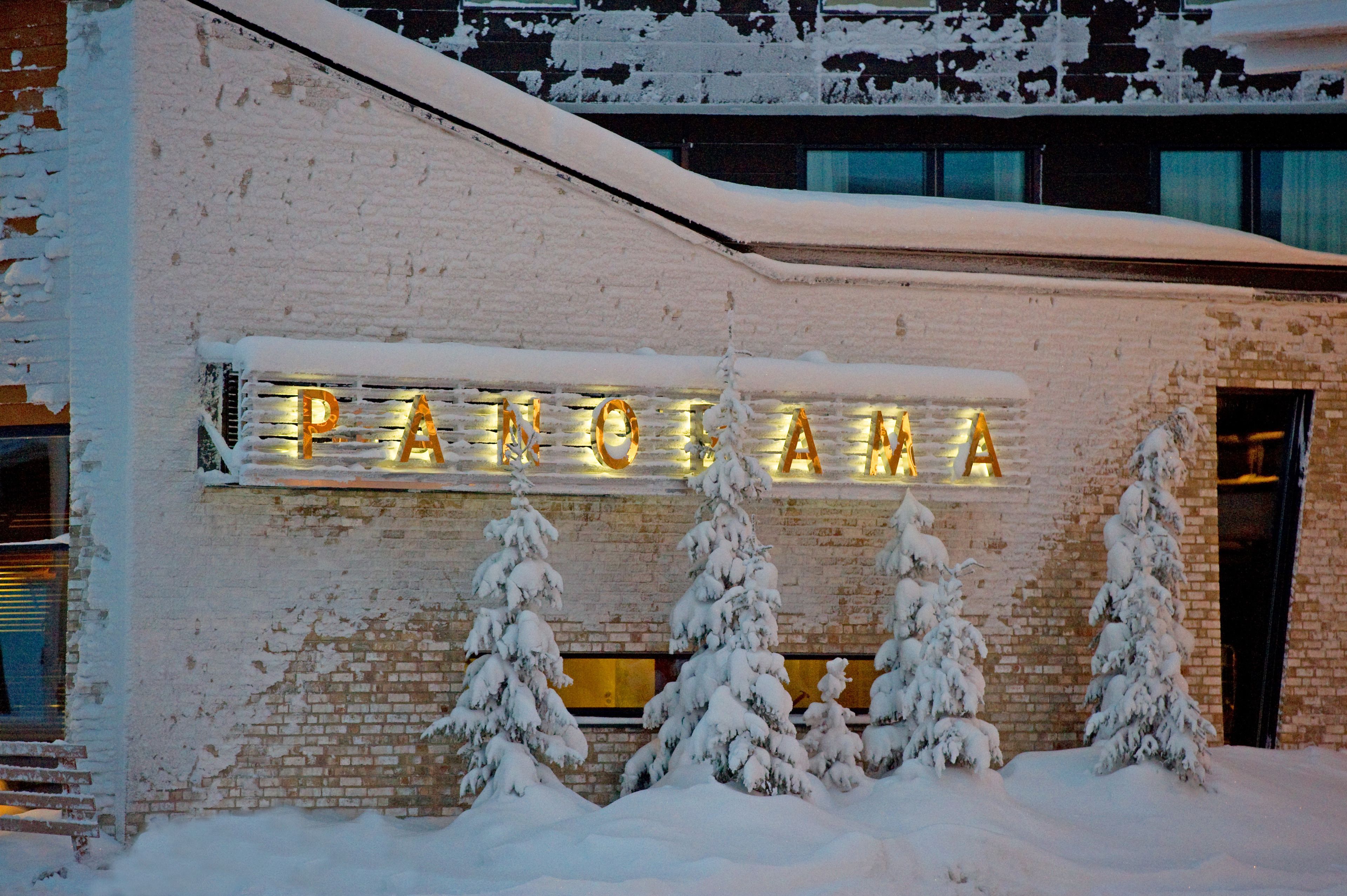 A building with a sign that reads "PANORAMA" illuminated in lights, surrounded by snow-covered trees.