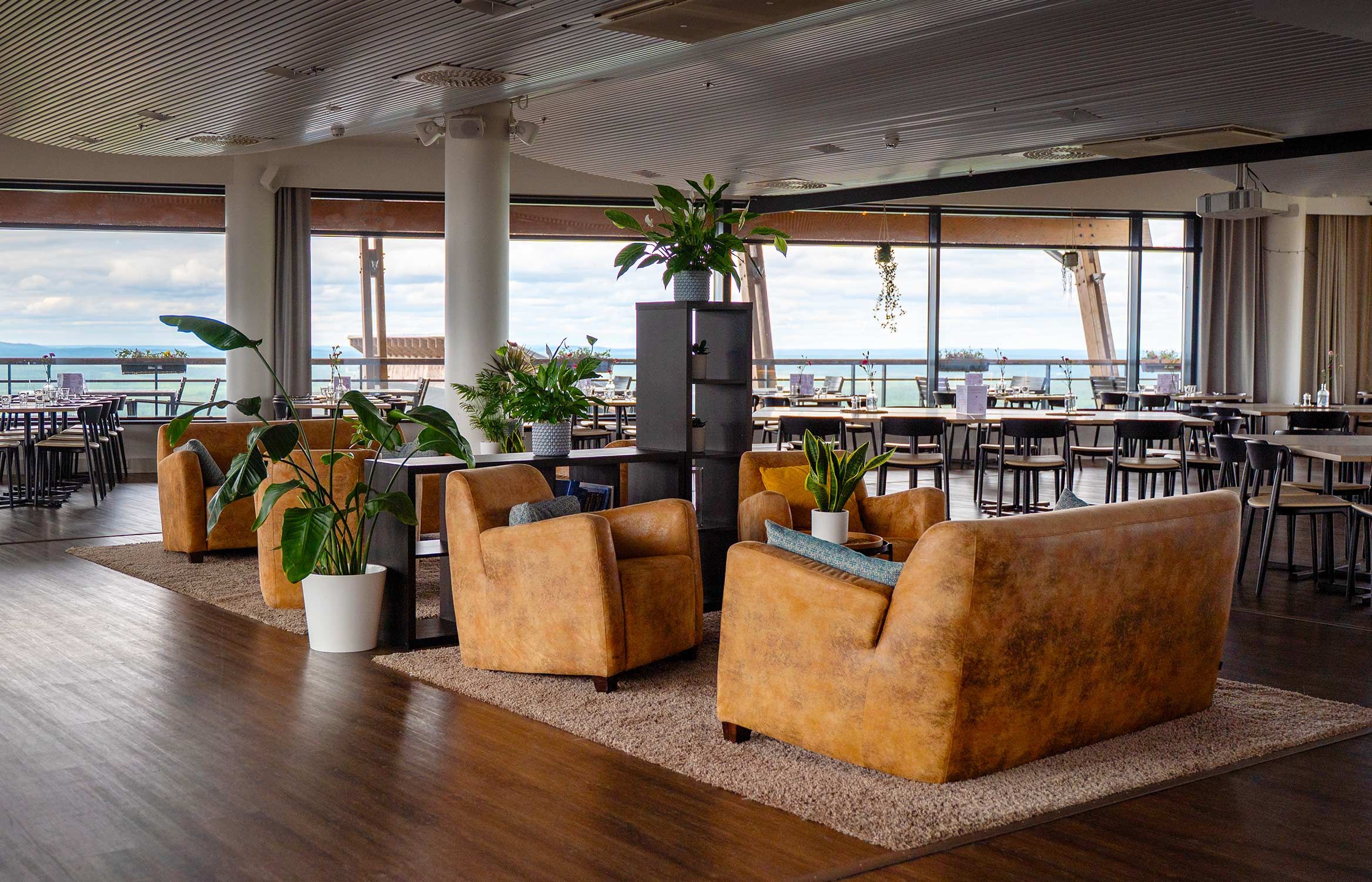 Amplia sala de estar con diversas opciones de asientos, incluyendo sofás y sillones, dispuestas alrededor de un pilar con plantas en macetas. Grandes ventanales ofrecen vistas al mar.